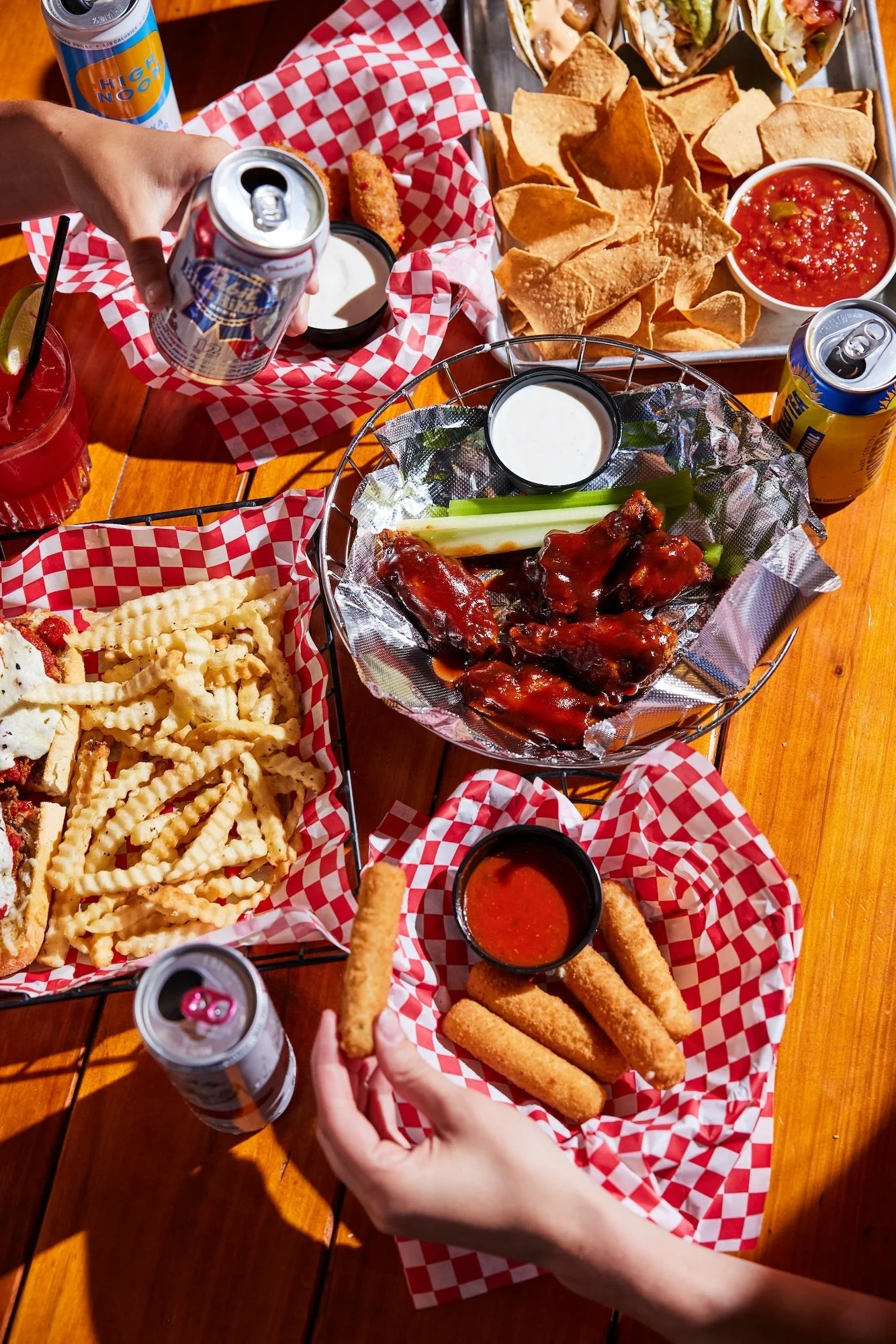 A table filled with various fried foods, chicken wings, cheese fries, chips with salsa, and drinks, with people reaching for the food.