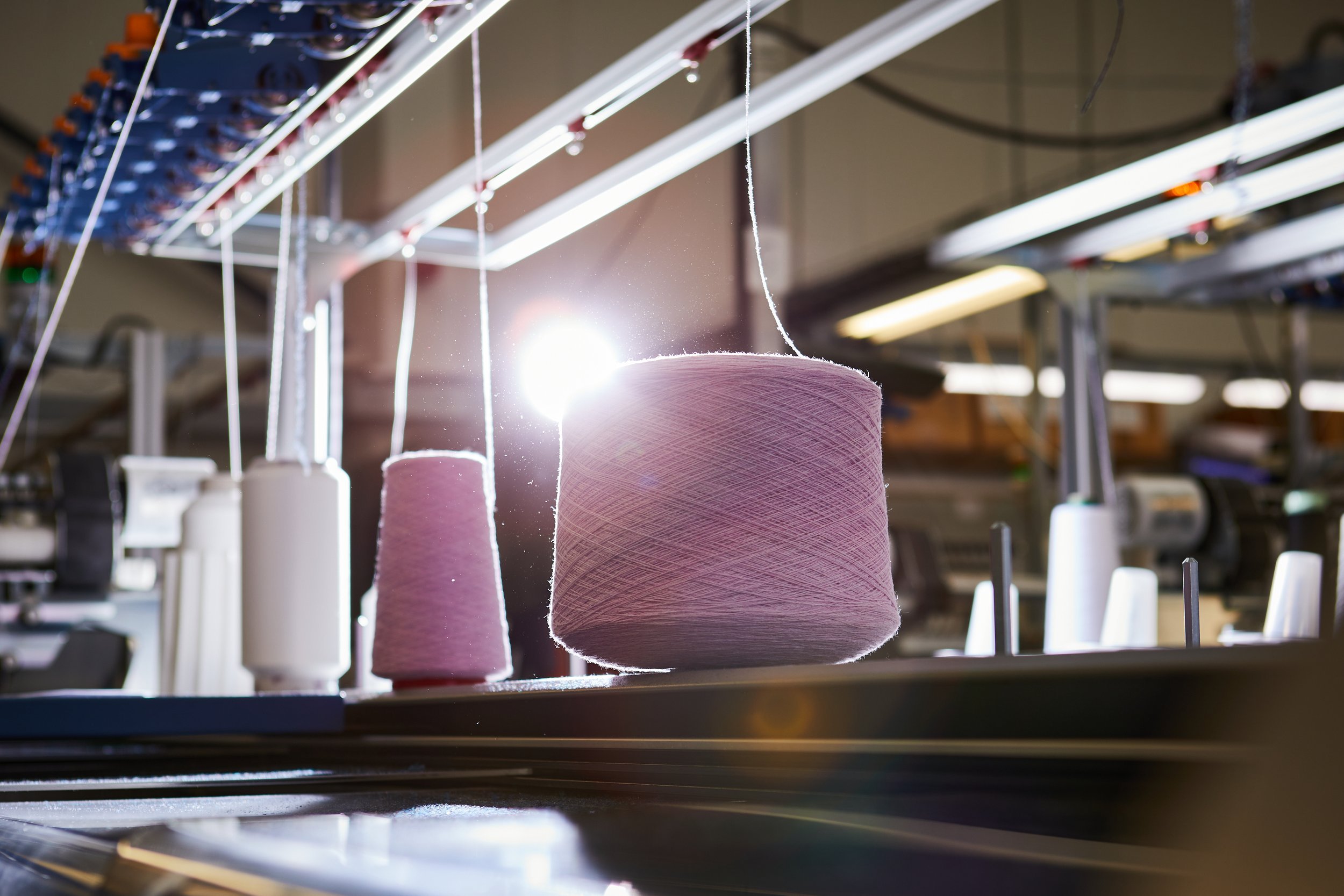Spools of pink and white thread hanging in a textile manufacturing or sewing factory, with sunlight shining through.