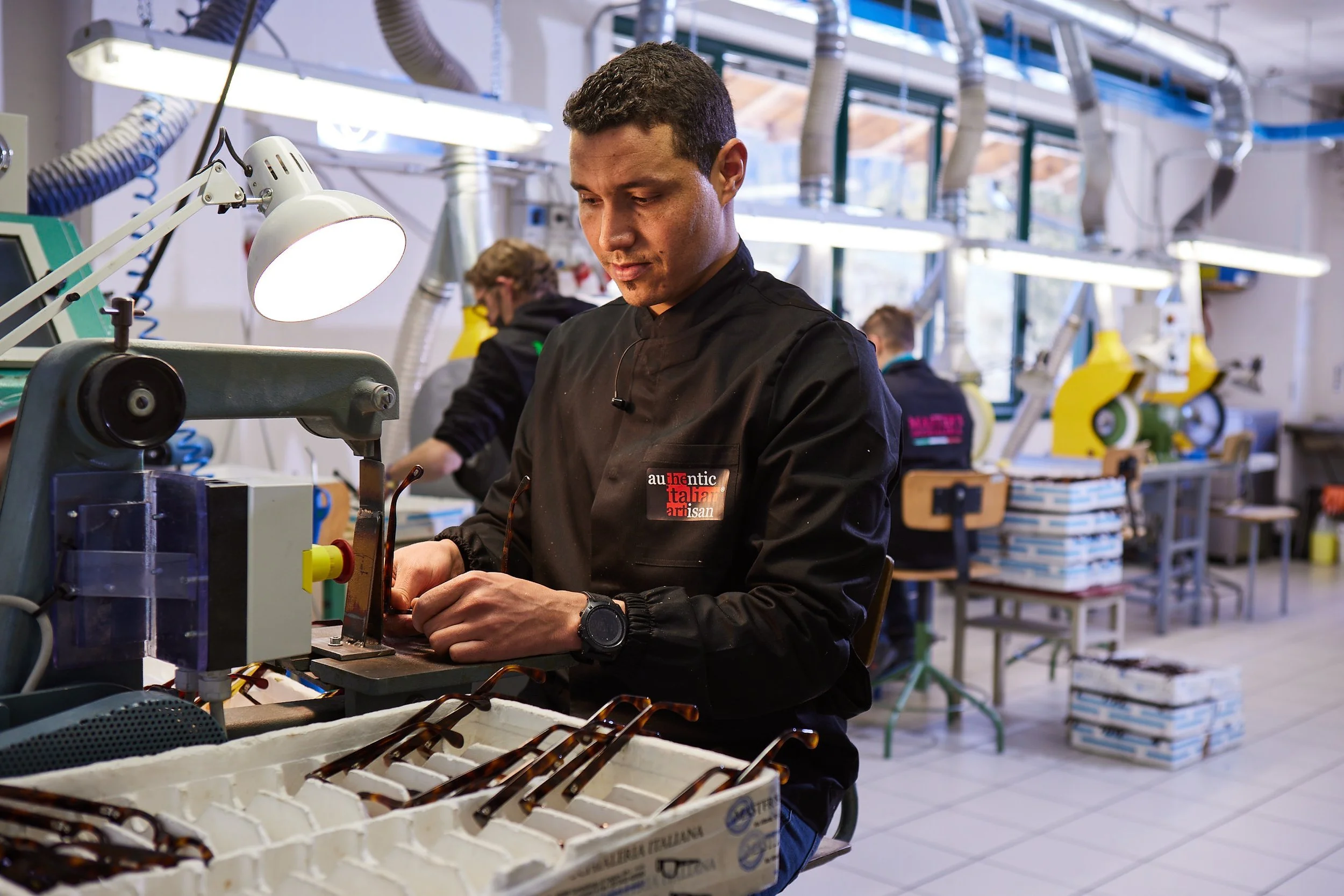 A man working on eyeglasses at a manufacturing or assembly workshop, with other workers and machinery visible in the background.