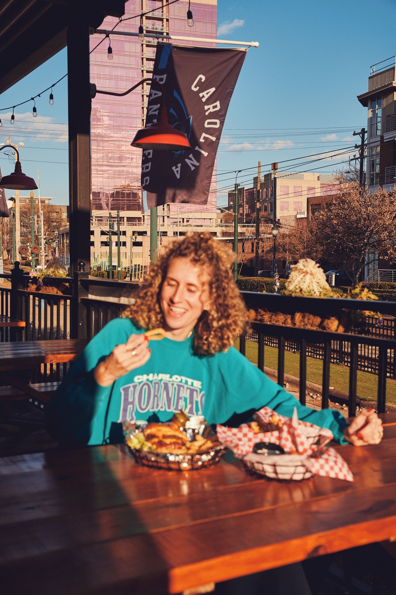 A woman with curly hair enjoying a meal at an outdoor restaurant or patio during late afternoon, with a Charlotte Hornets sweatshirt, a flag with a sports team logo, and buildings in the background.