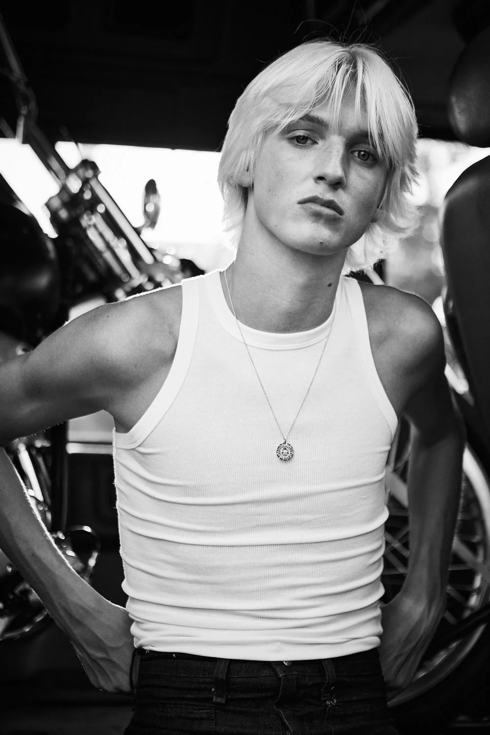 Black and white photo of a young person with short, platinum blonde hair wearing a white tank top and a pendant necklace, standing next to a motorcycle.