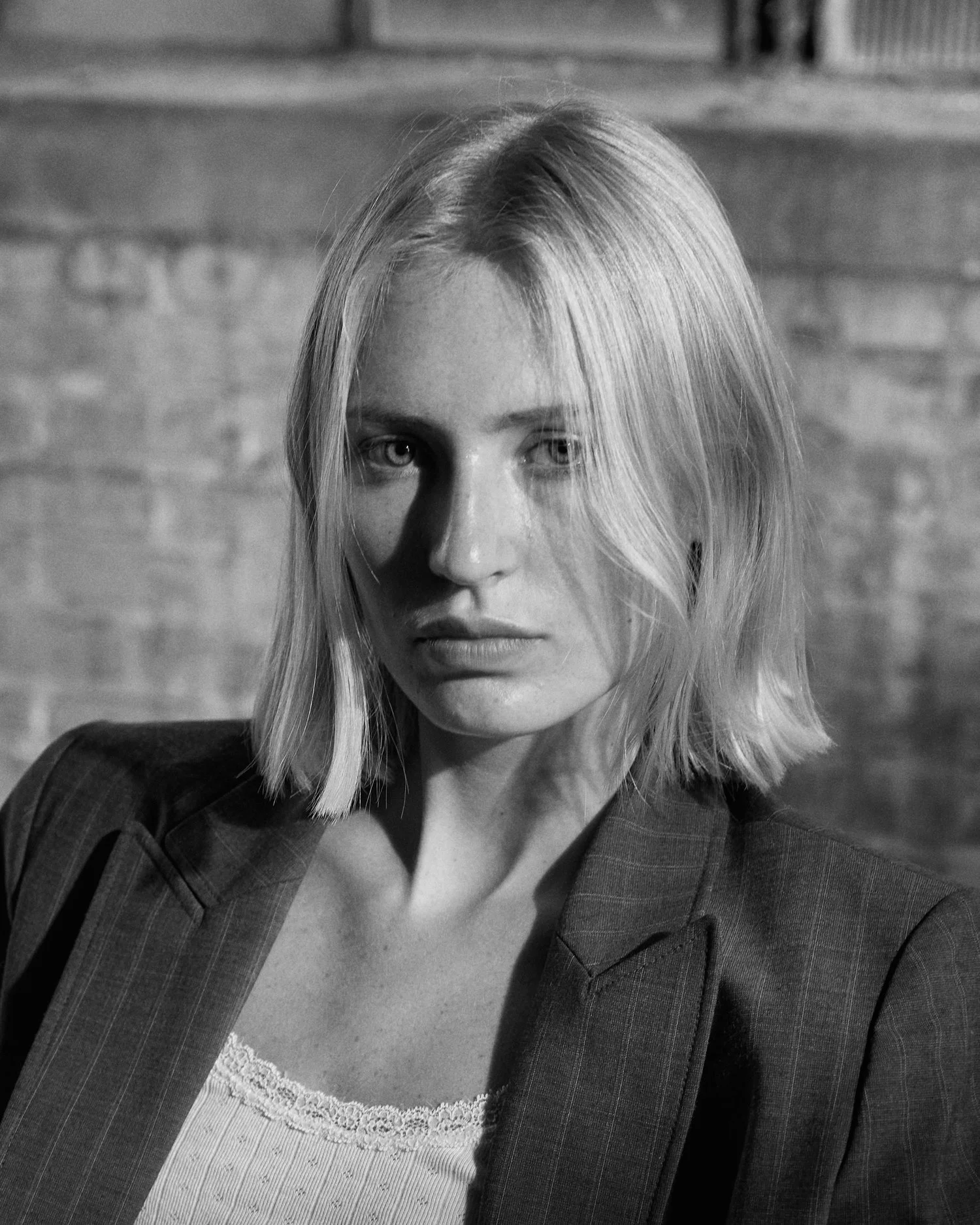 Black and white portrait of a woman with shoulder-length blonde hair, wearing a jacket over a lace-trimmed top, standing against a brick wall.