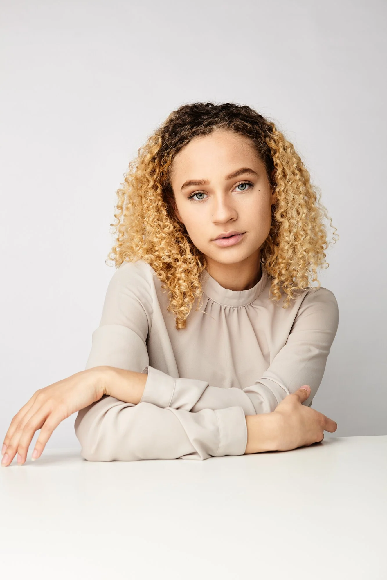 Young woman with curly blonde hair sitting at a white table, wearing a beige long-sleeve top, looking at the camera with a neutral expression.