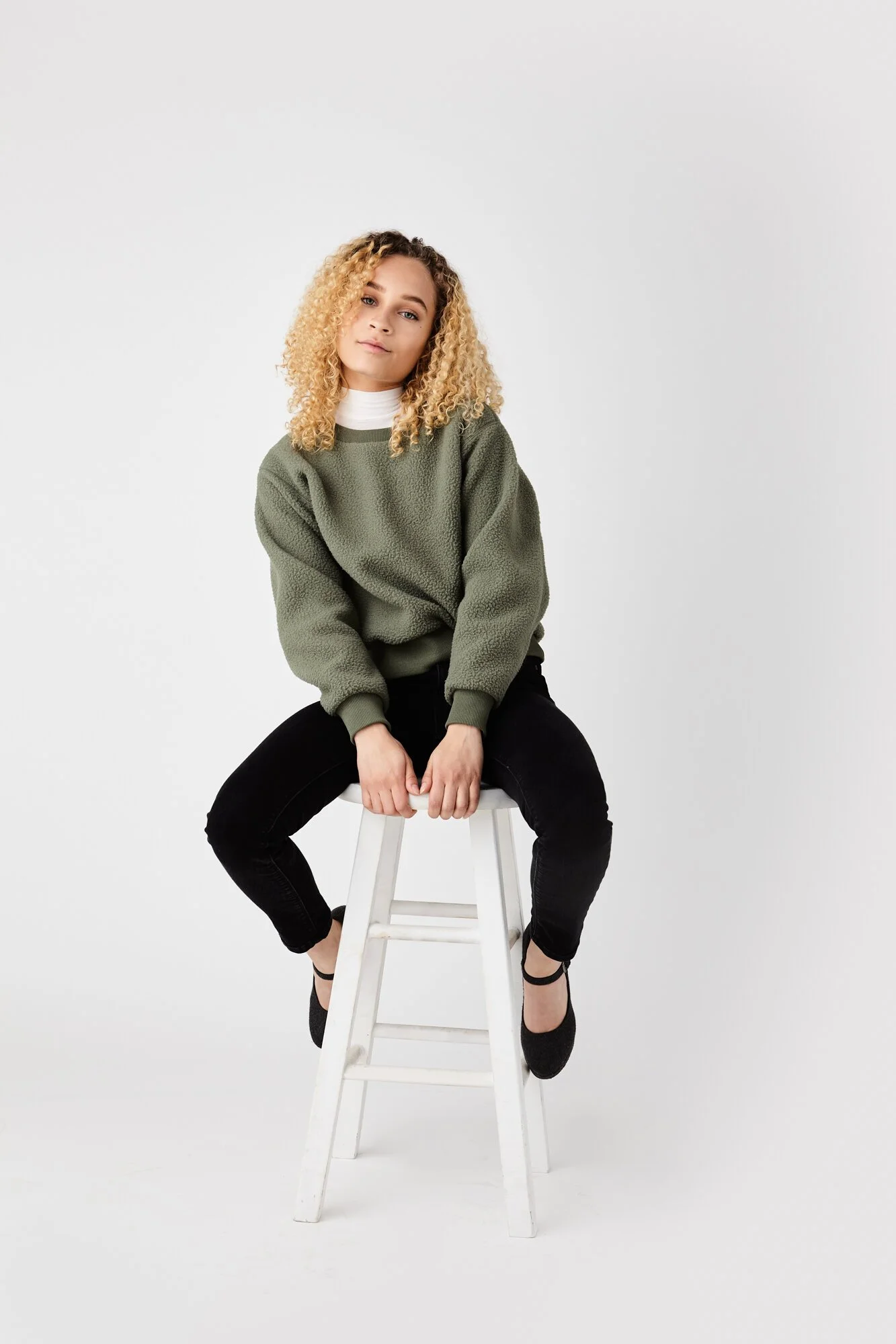 A young woman with curly blonde hair sitting on a white stool against a plain white background. She is wearing a green fleece sweater, black pants, and black shoes.