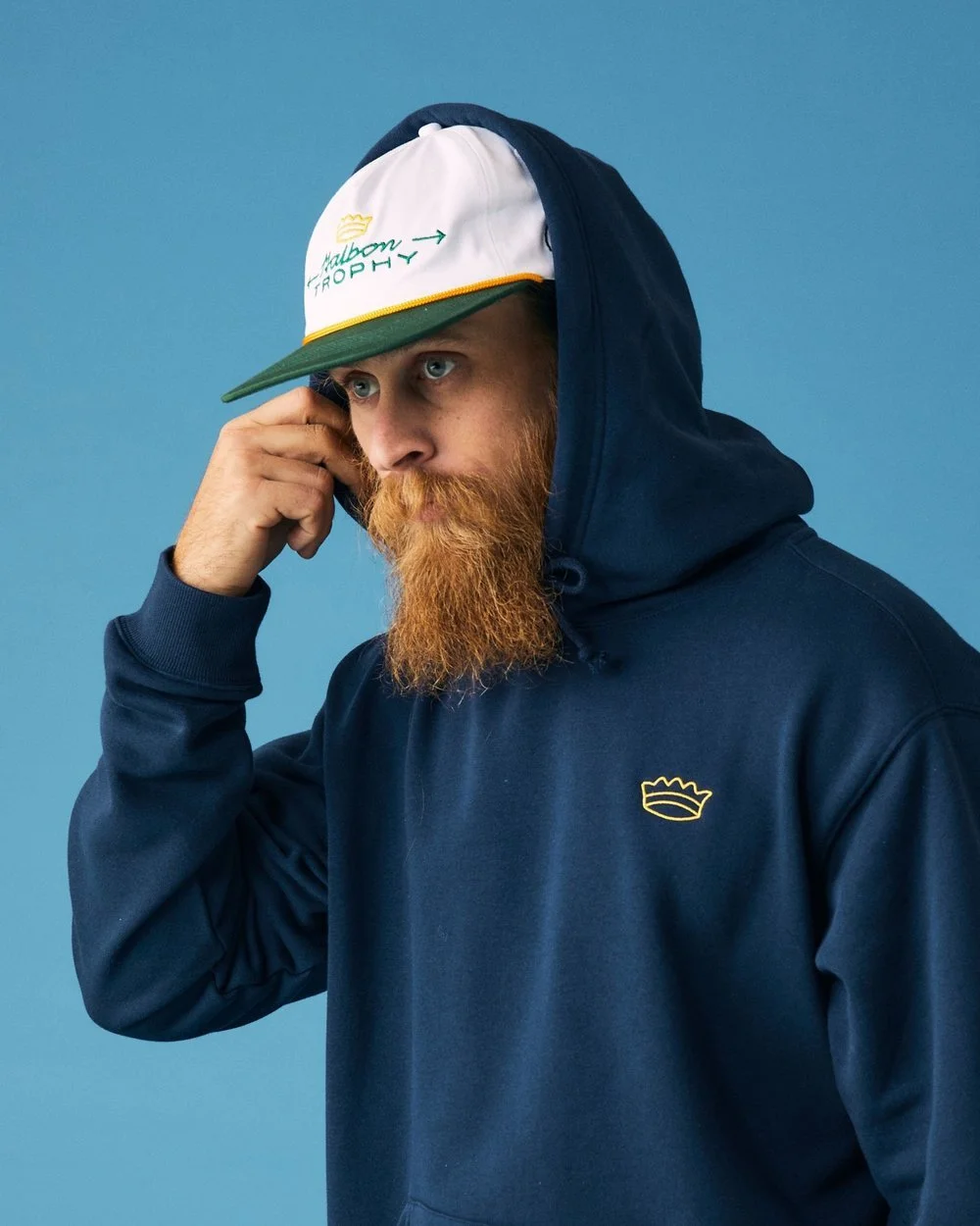 A man with a red beard wearing a navy blue hoodie and a colorful cap, touching his temple, against a blue background.