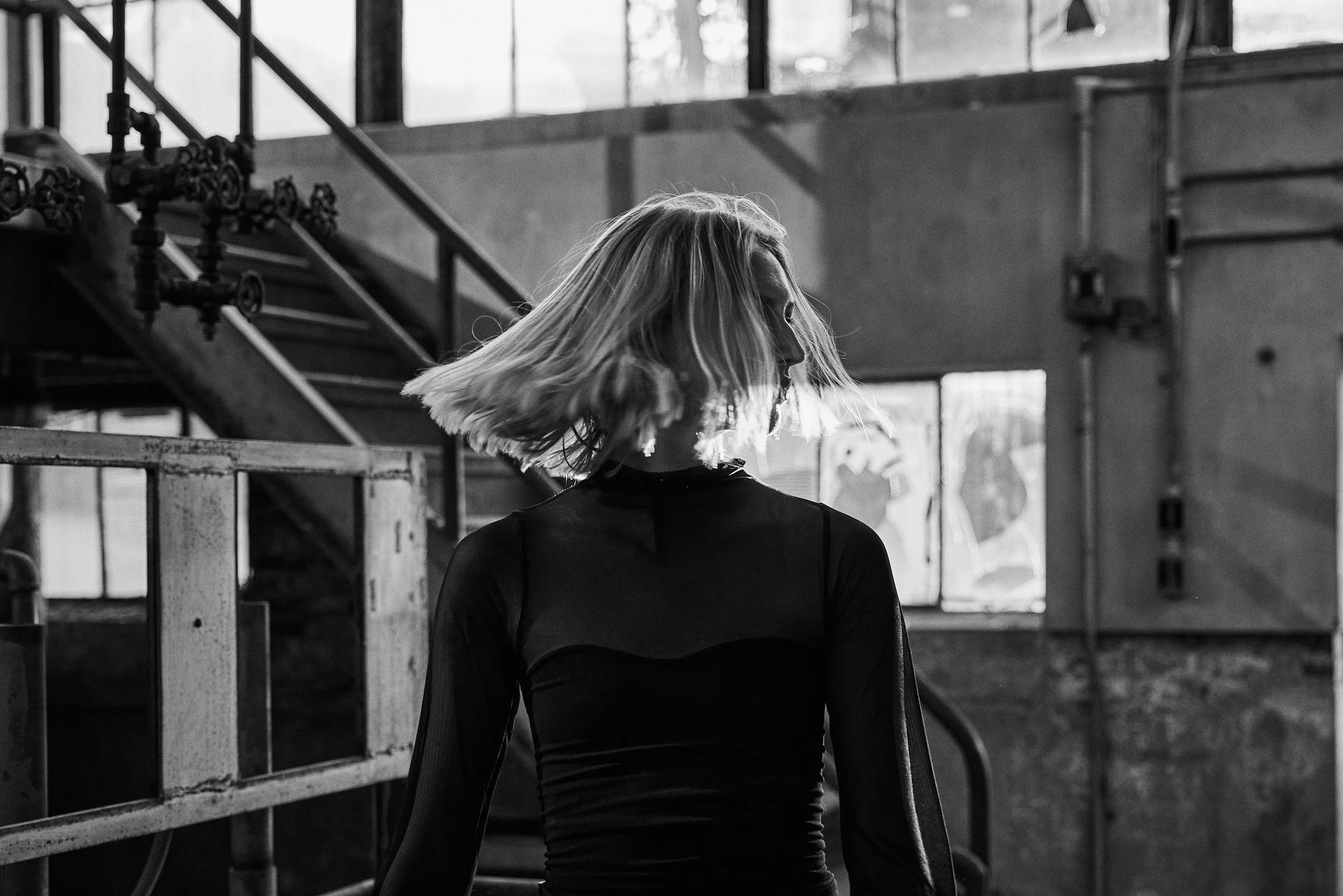 A woman with shoulder-length blonde hair, wearing a black long-sleeve top, is spinning or moving in an industrial or warehouse-like space with metal railings and stairs in the background. The photo is in black and white.
