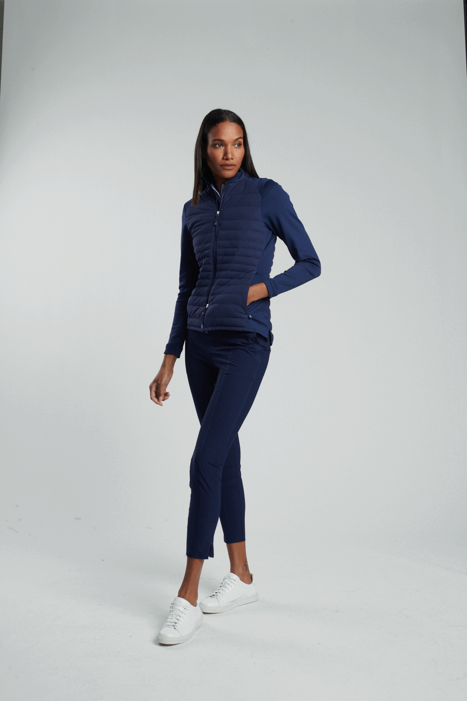 A woman wearing a navy blue quilted jacket, navy blue pants, and white sneakers poses against a plain light gray background.
