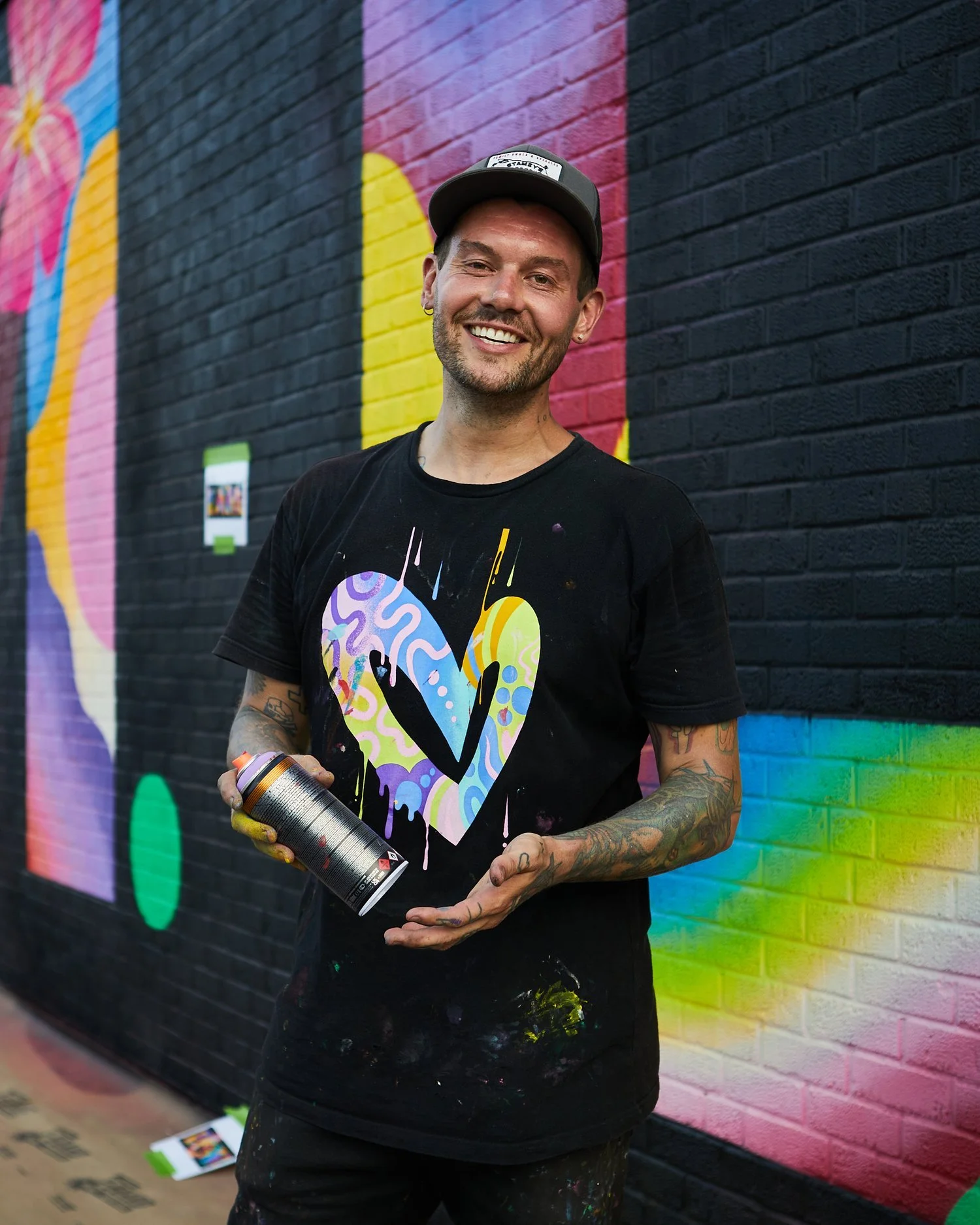 Male street artist smiling while holding a spray paint can, standing in front of a colorful mural on a brick wall.