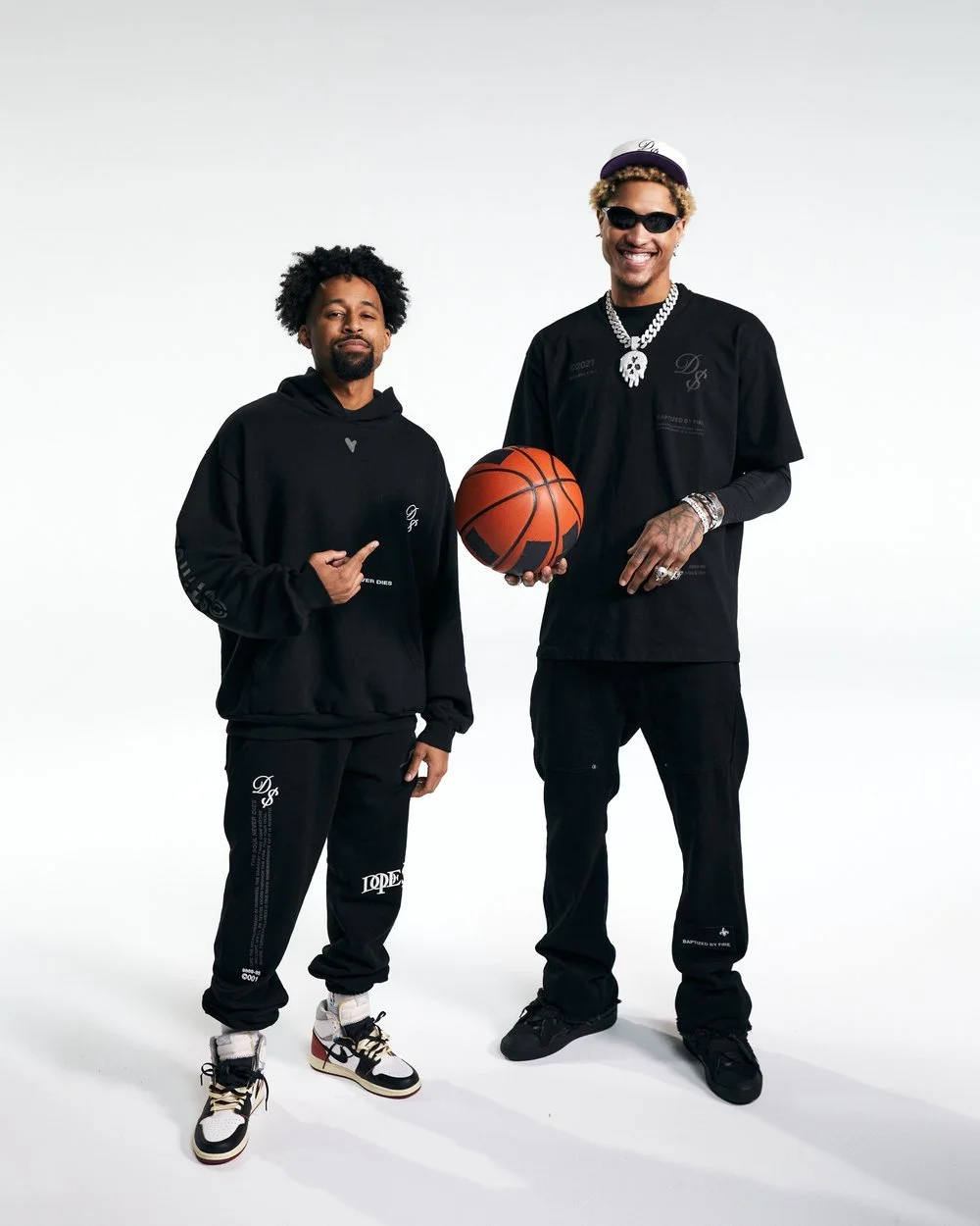 Two men dressed in black clothing, one holding a basketball, standing against a plain white background, smiling for the camera.