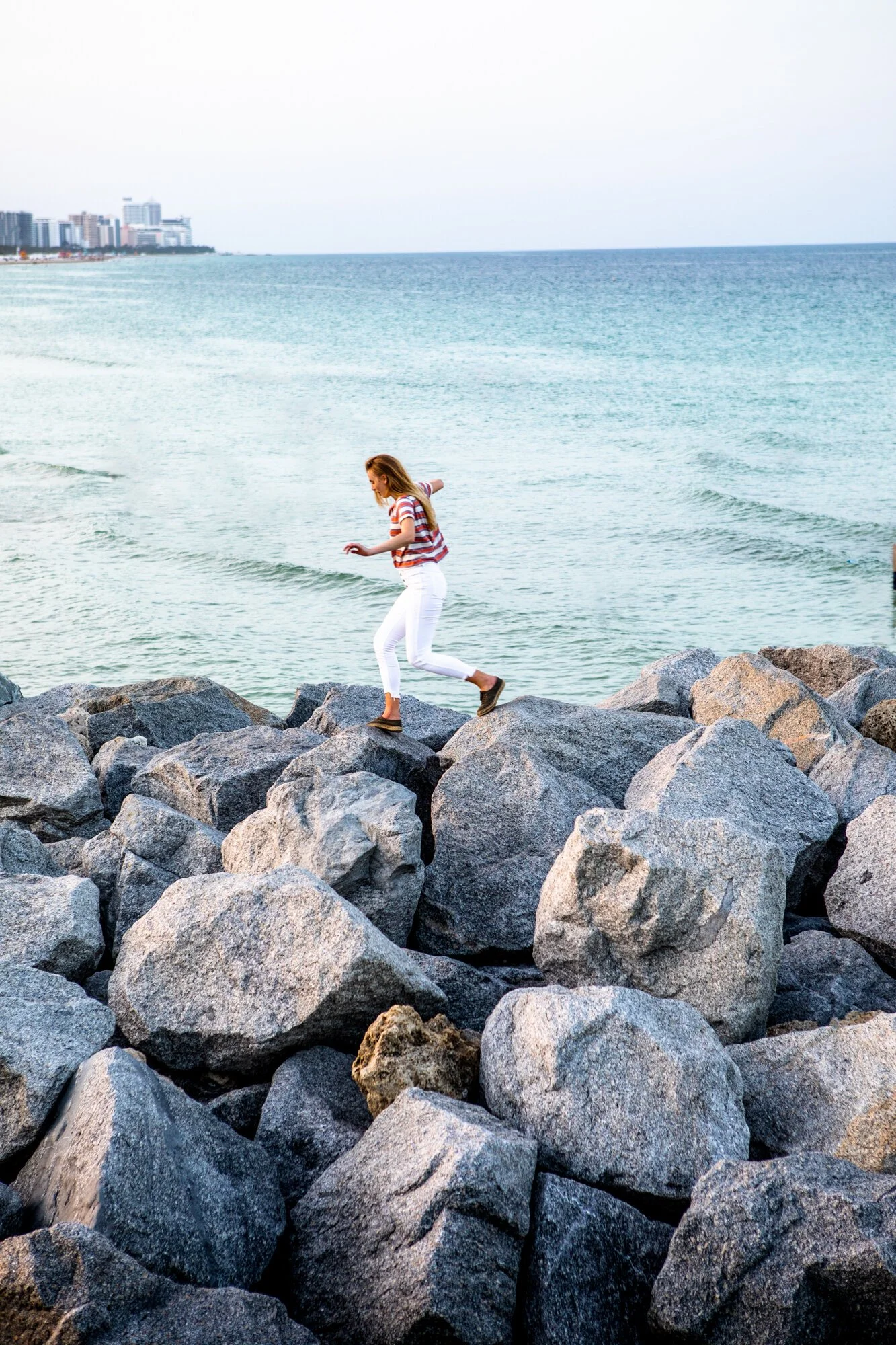 A woman with red hair, wearing a striped shirt and white pants, balancing on large rocks by the ocean near a city skyline.