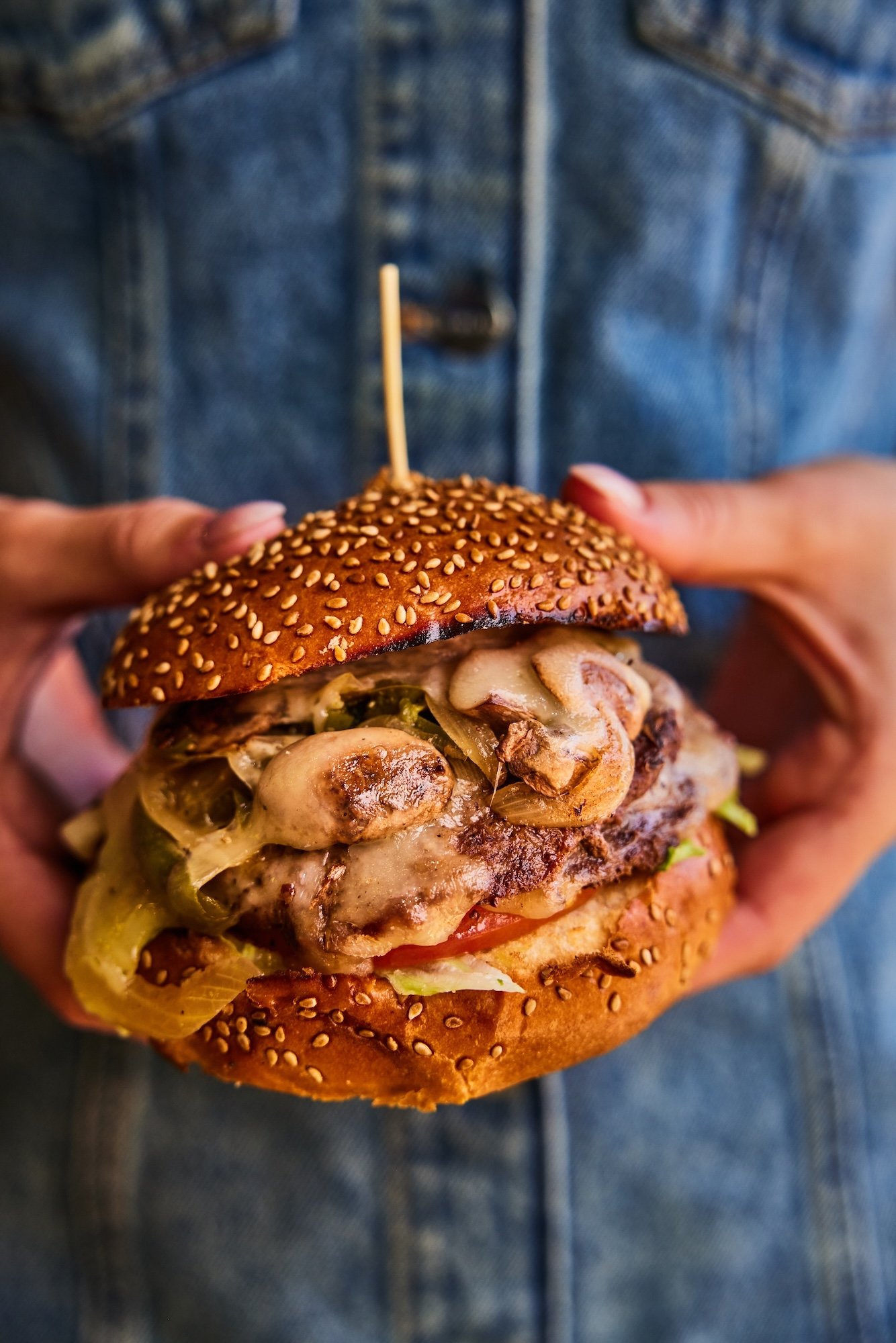 Person holding a large cheeseburger with melted cheese, grilled beef patty, lettuce, tomato, pickles, and sesame seed bun.