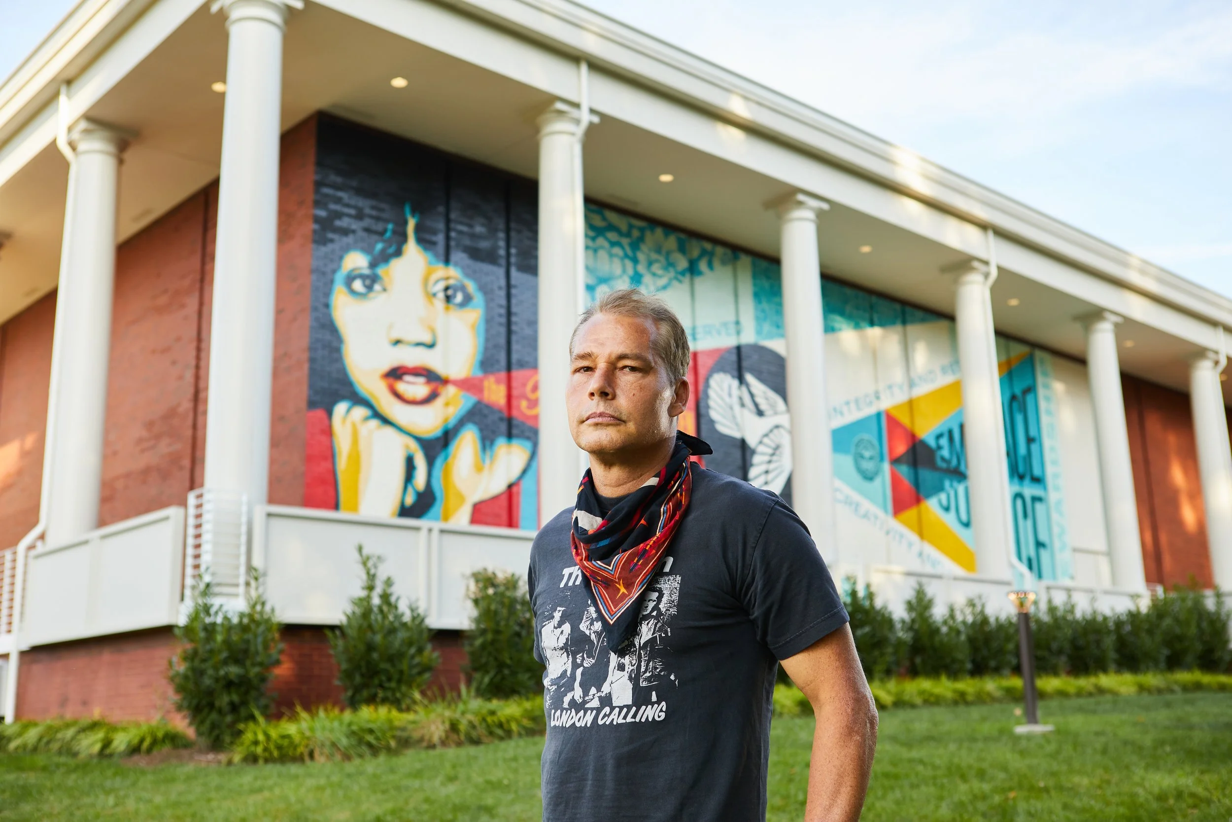 A man standing in front of a building with large colorful murals, one depicting a woman's face with a cigarette, and another with geometric shapes and text.