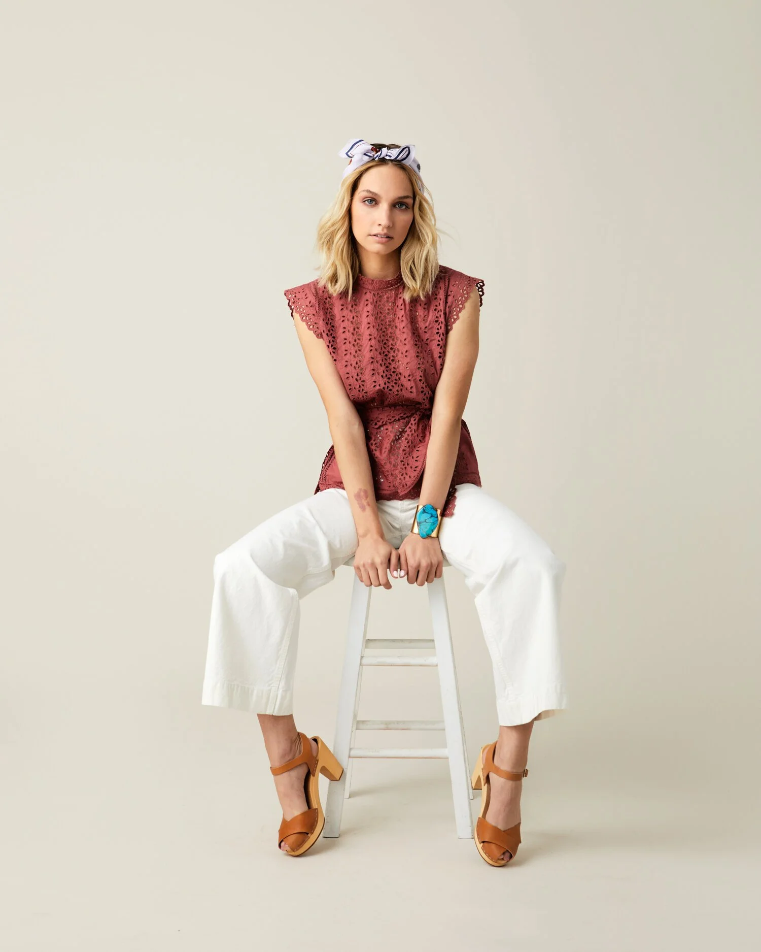 Young woman with blonde hair wearing a white headscarf with navy blue patterns, a red lace sleeveless top, white wide-leg pants, and tan high heels, sitting on a white stool against a plain light background.