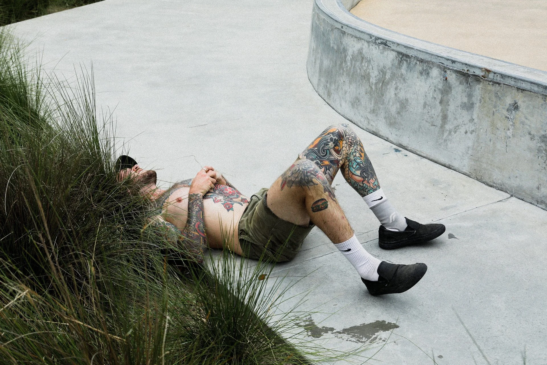 A tattooed man lying on the ground next to some grass, resting with eyes closed, wearing sunglasses, a cap, shorts, white Nike socks, and black shoes, near a concrete skatepark ramp.