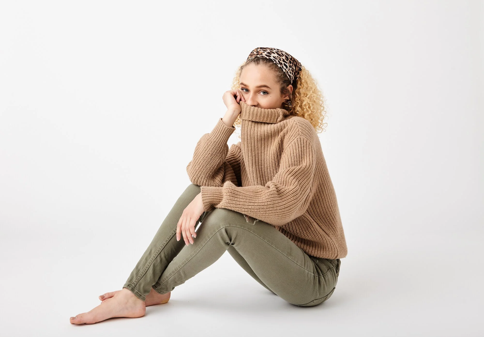 Young woman with curly blonde hair wearing a leopard print headband and tan chunky knit sweater, sitting on the floor against a plain white background, partially covering her face with her sweater as she looks at the camera.