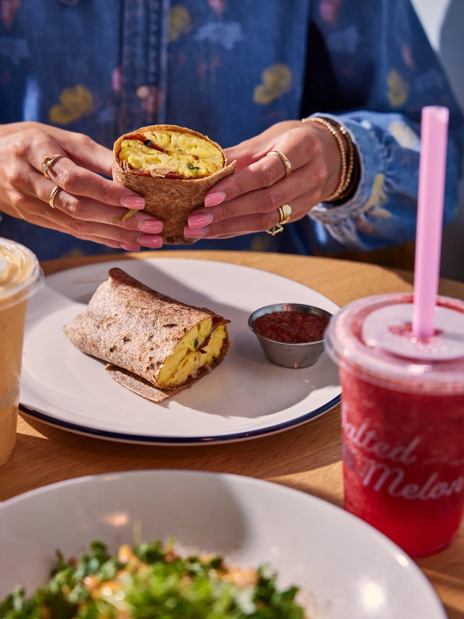 Person holding a slice of breakfast burrito with scrambled eggs, cheese, and potatoes. On the table, there is a rolled breakfast burrito, a small container of salsa, a pink drink with a straw, and other dishes.