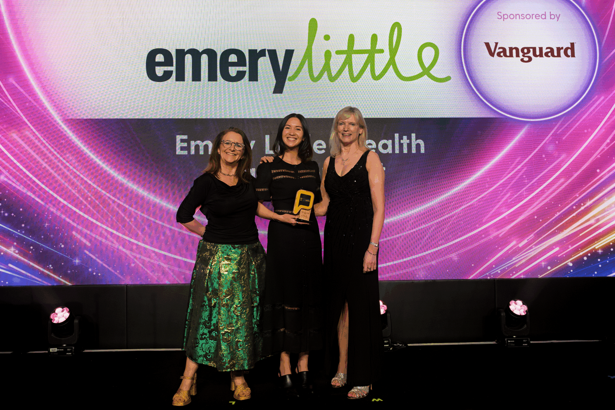 Three people standing side-by-side  on stage facing towards us. One person in the centre is holding an award. The group is standing in front of a screen displaying the Emery Little logo, sponsored by Vanguard, at the 2026 Professional Adviser Awards.