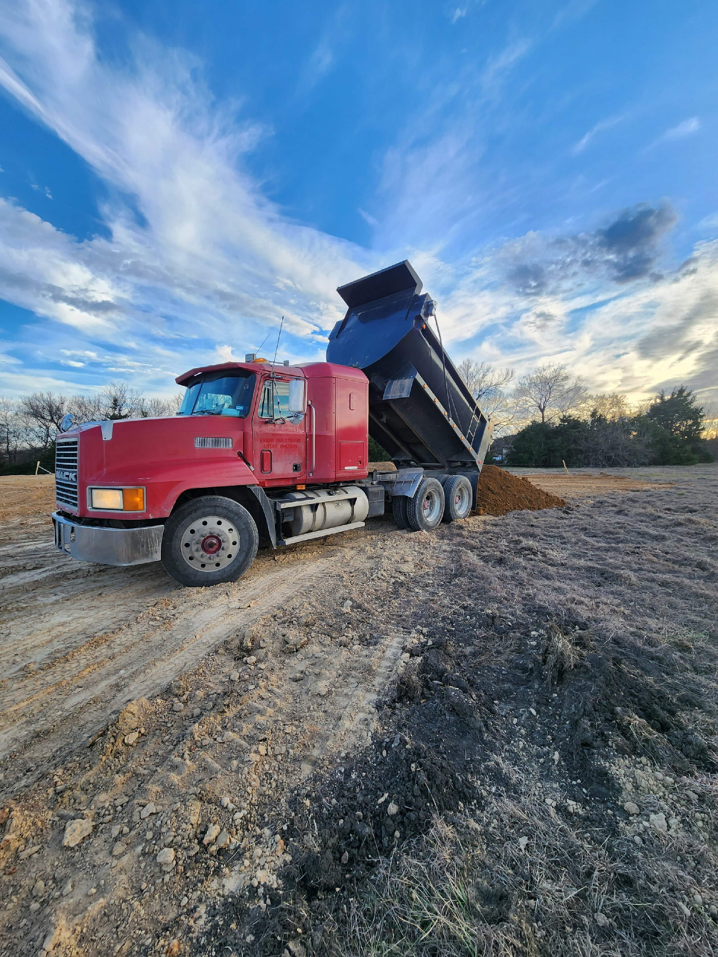 A red dump truck unloading dirt on a construction site with a partly cloudy sky in the background.