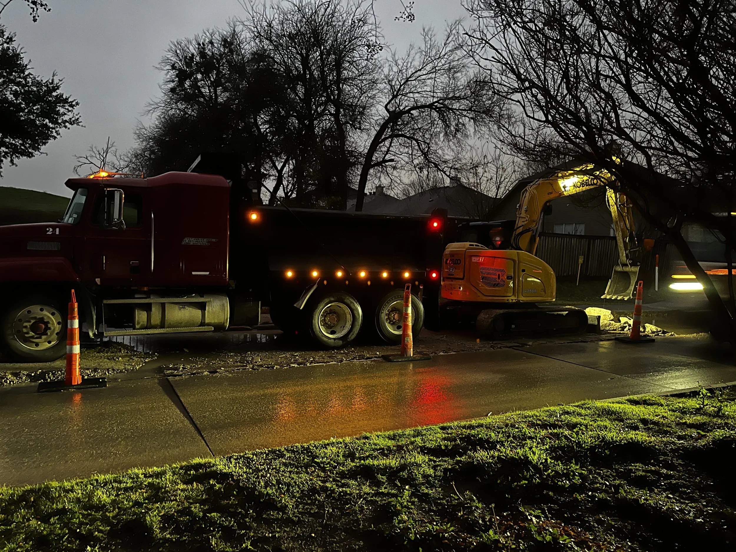 A construction site on a rainy evening with a red dump truck and a yellow mini excavator surrounded by orange traffic cones, with wet pavement reflecting the lights and leafless trees in the background.