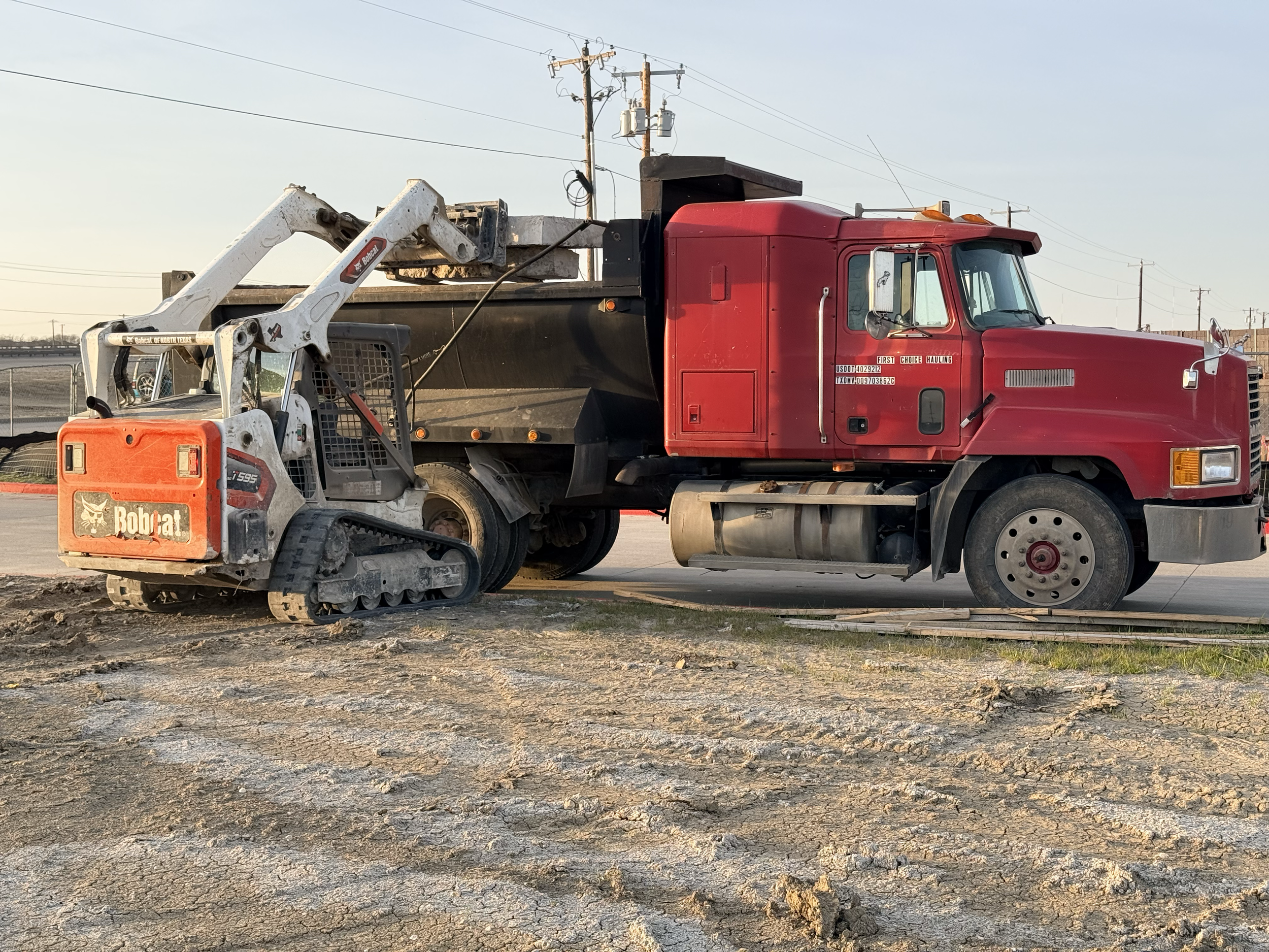 Red dump truck and Bobcat construction vehicle on a construction site with dirt and gravel ground.