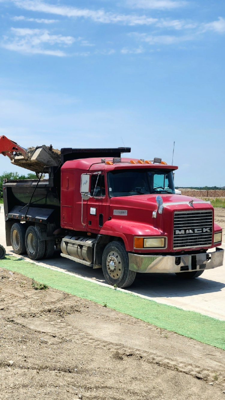 Red dump truck with a loader bucket on a construction site, under a cloudy blue sky.
