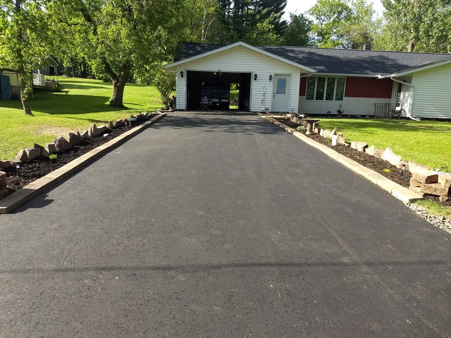 A freshly paved driveway leading to a garage attached to a white house, with landscaped borders featuring rocks and small plants on either side, green lawn, trees, and a neighboring house visible in the background.