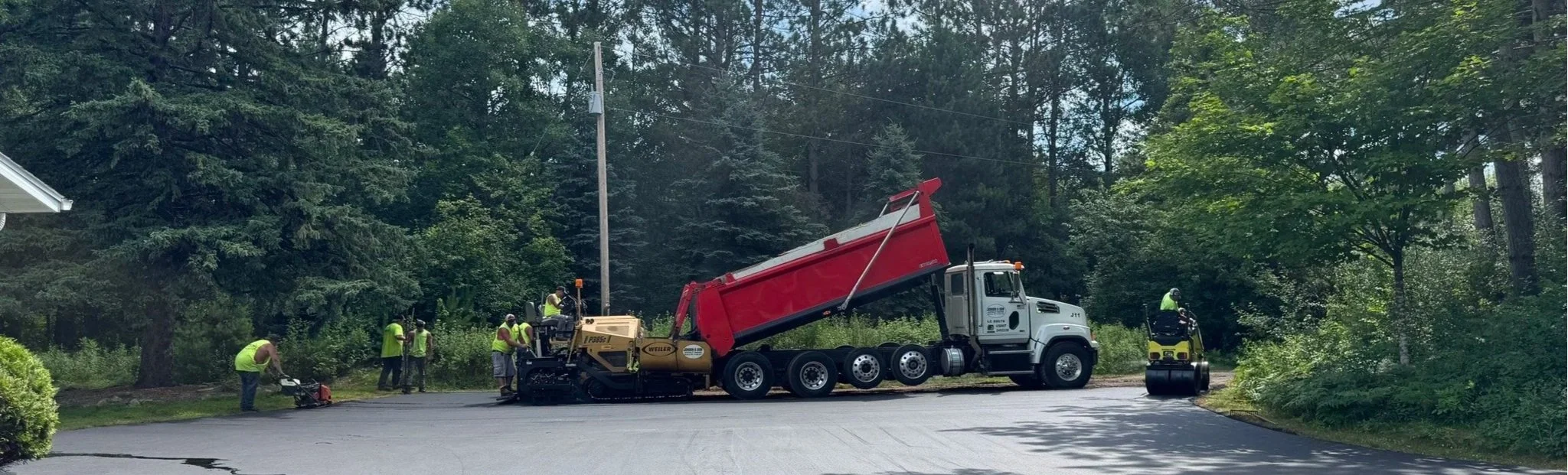 Asphalt pavers in yellow safety vests working on a residential Wisconsin driveway near a wooded area, with a large red and white dump truck and various machinery.