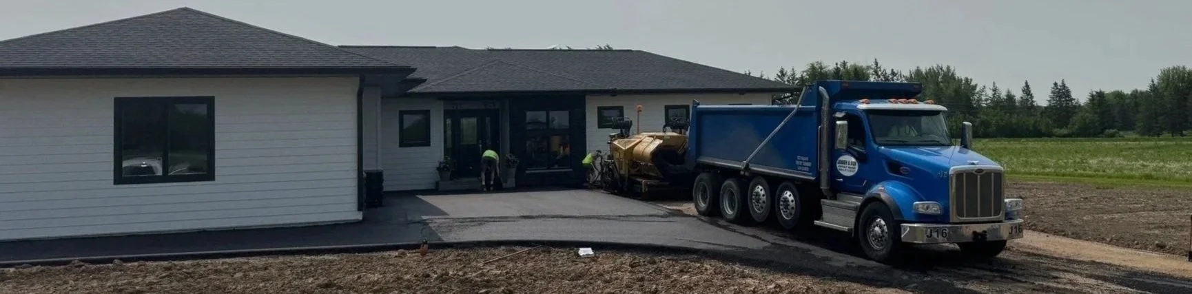 A residential Wisconsin house with a new driveway being poured. Asphalt pavers are working near the entrance. A large blue dump truck is parked on the working driveway.