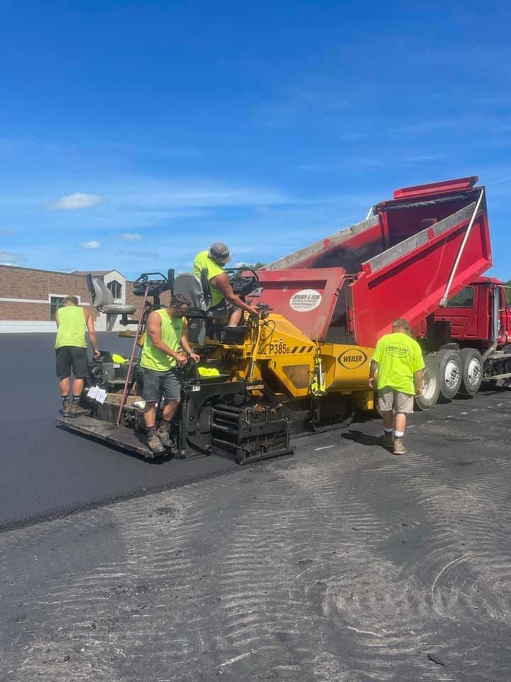 Workers in neon shirts paving a new asphalt road with a paving machine under a clear blue sky.