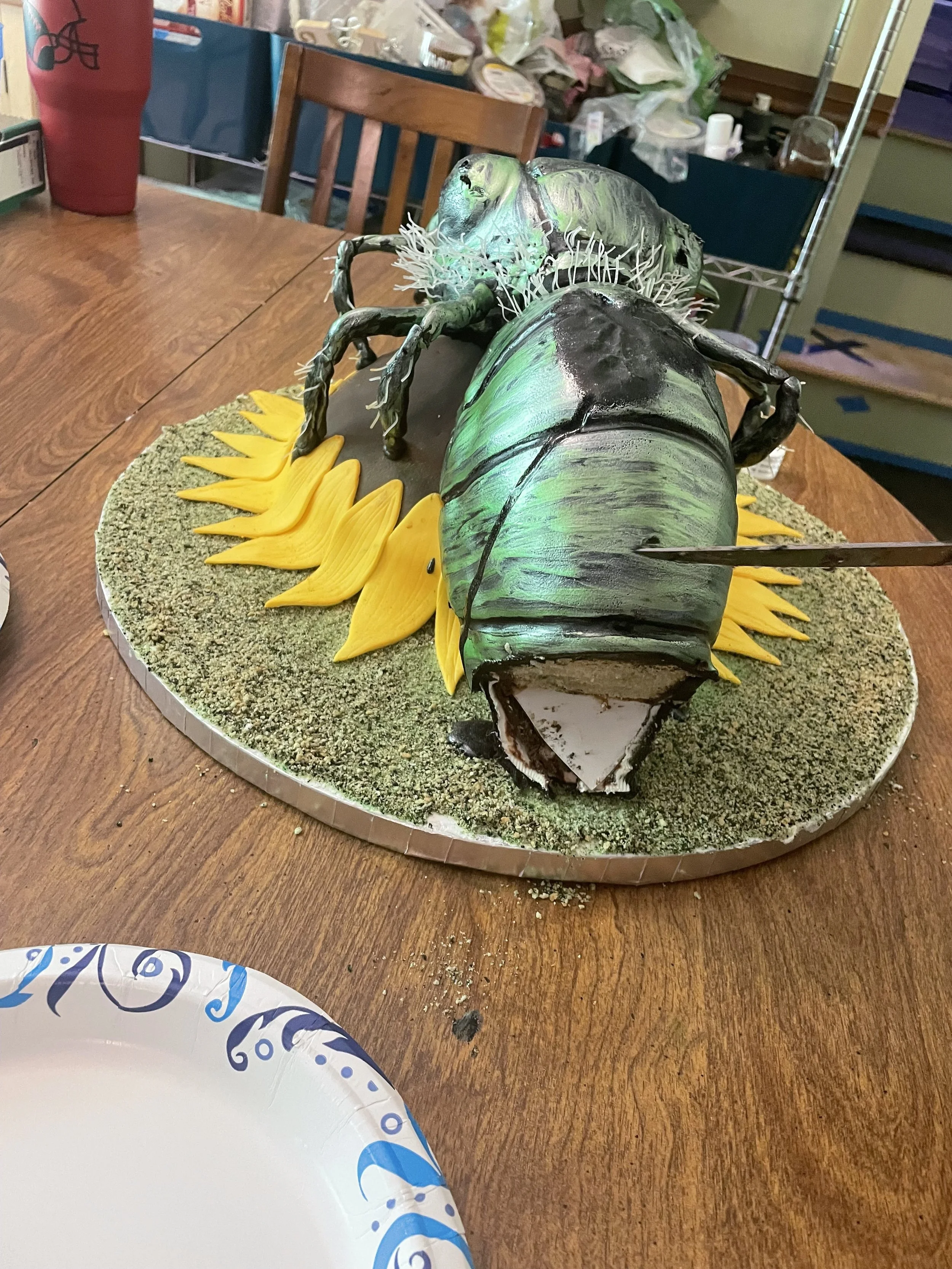 A cake designed to look like a large green and black insect, resting on a base of sunflower petals and green textured ground. The cake features detailed edible decorations, including the insect's body, wings, and legs, displayed on a wooden table wit