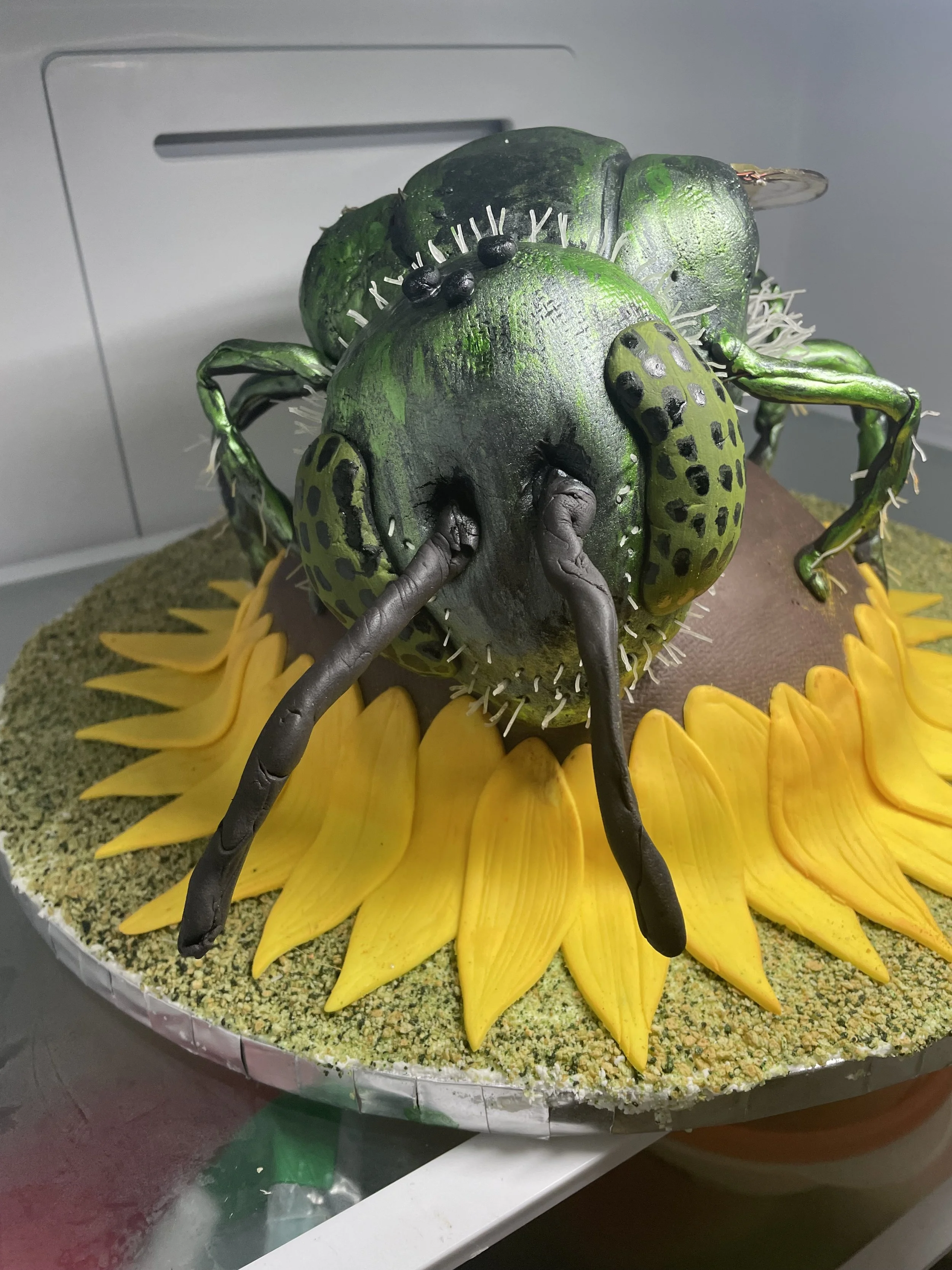Cake decorated as a large, green insect, green metallic sweat bee, with yellow petal-like decorations around the base, on a circular platform.