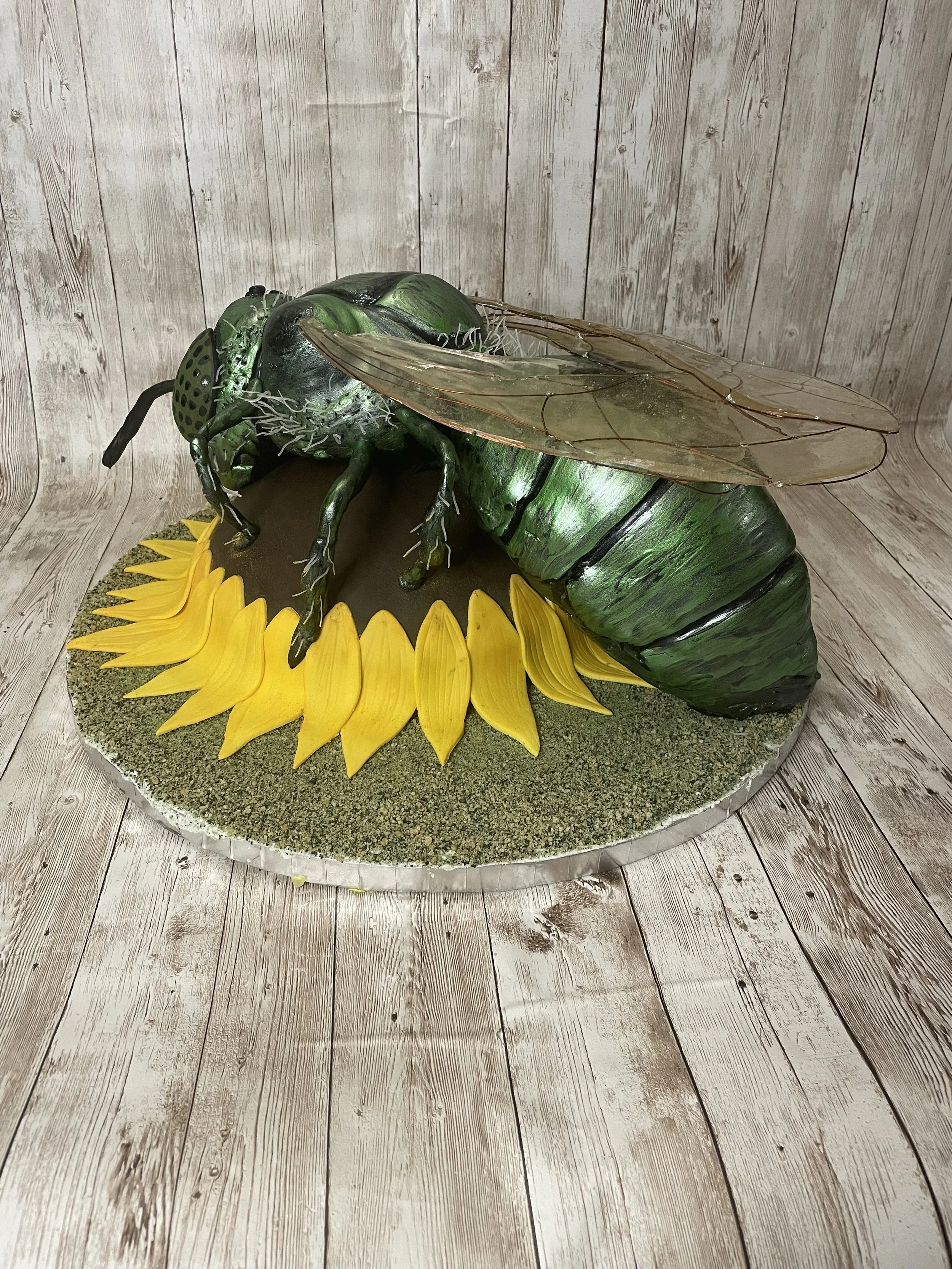 A cake designed to look like a realistic green metallic sweat bee insect on a yellow sunflower base, with detailed wings, legs, and antennae, set on a round cake board against a wood-paneled background.