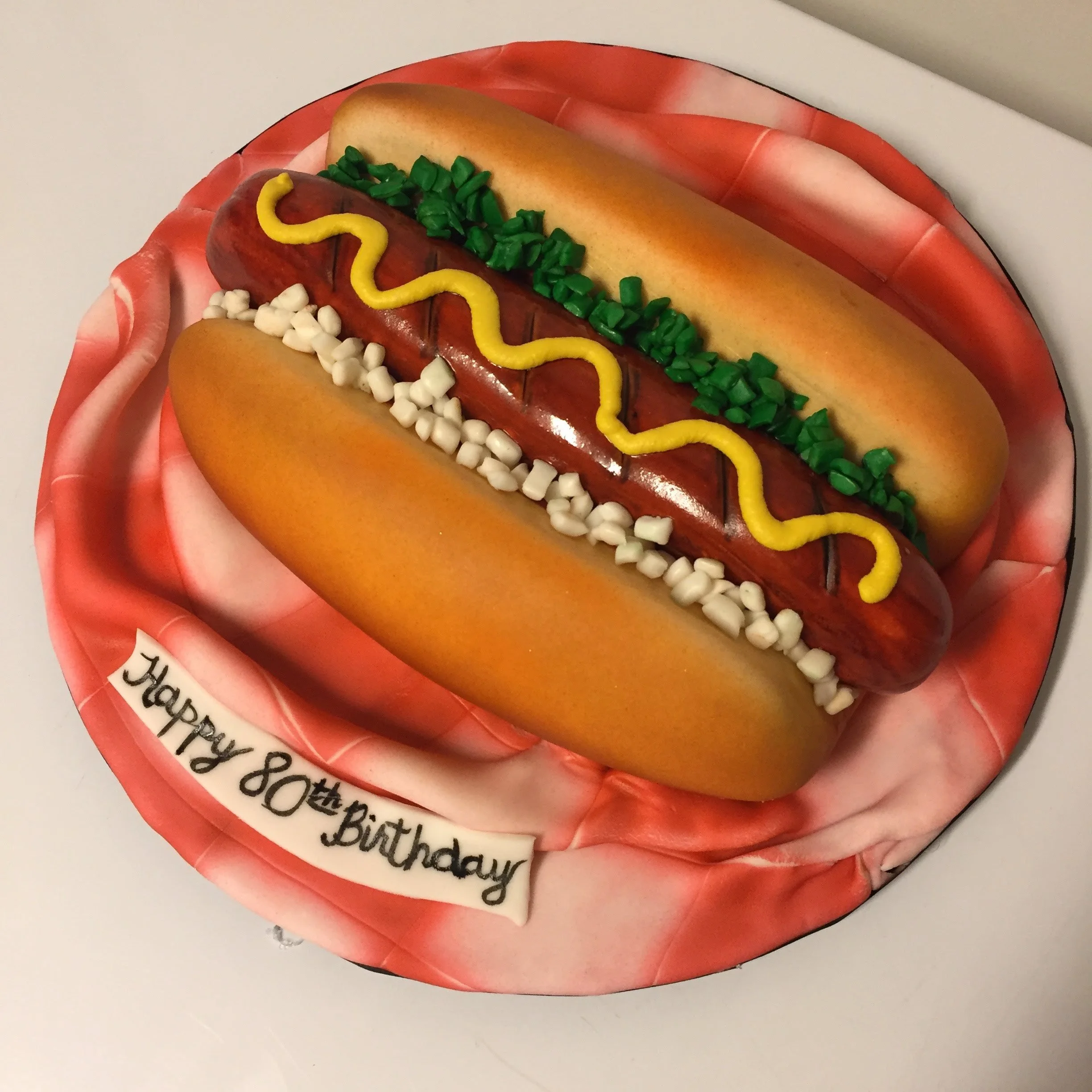 A cake shaped like a hot dogs with a bun, toppings, and mustard, on a plate with a birthday message.