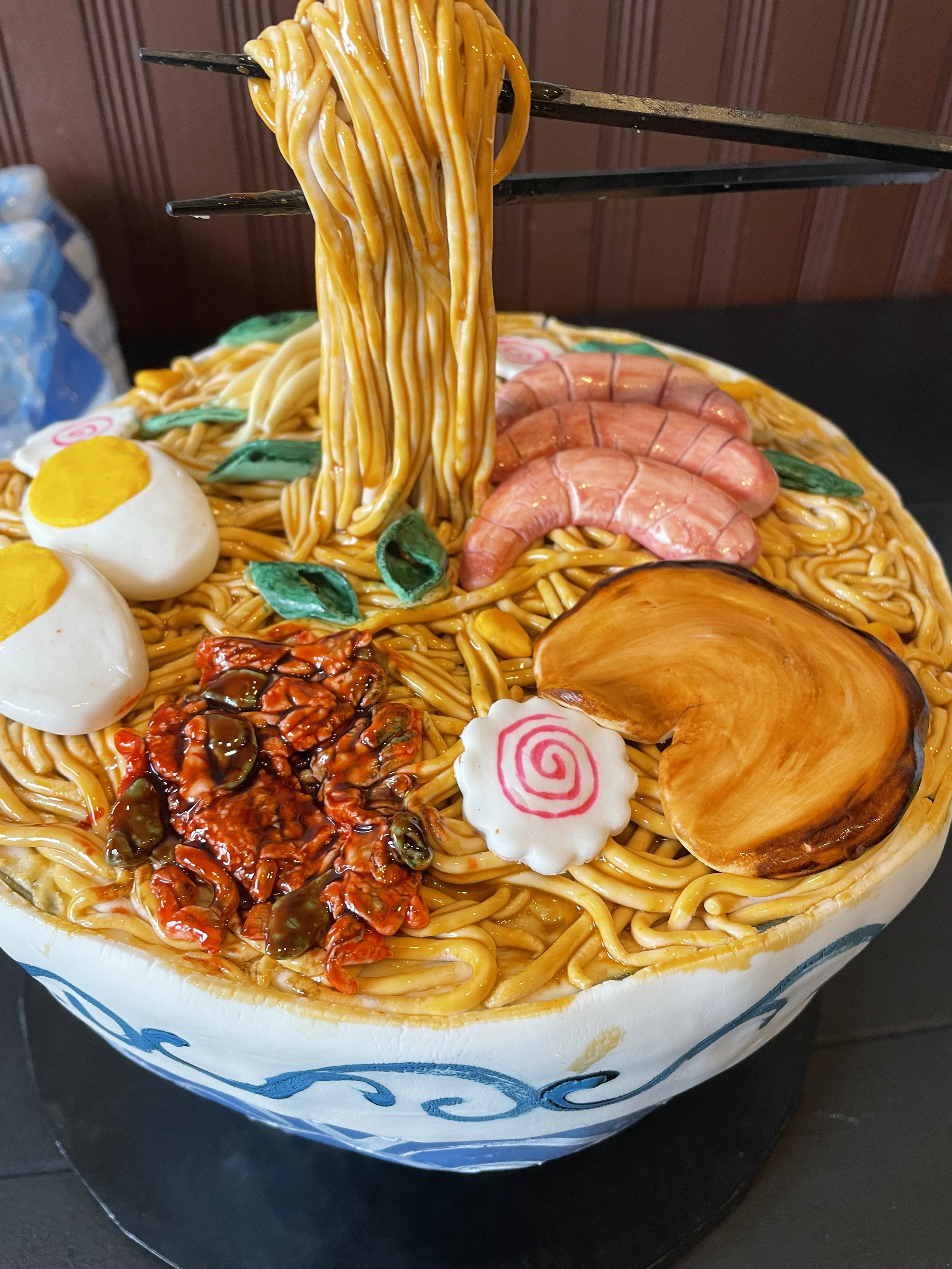 A large bowl of ramen noodles cake topped with various food items like boiled eggs, shrimp, sliced meat, narutomaki, sliced pumpkin, and spicy chili sauce, with noodles being lifted by black chopsticks.