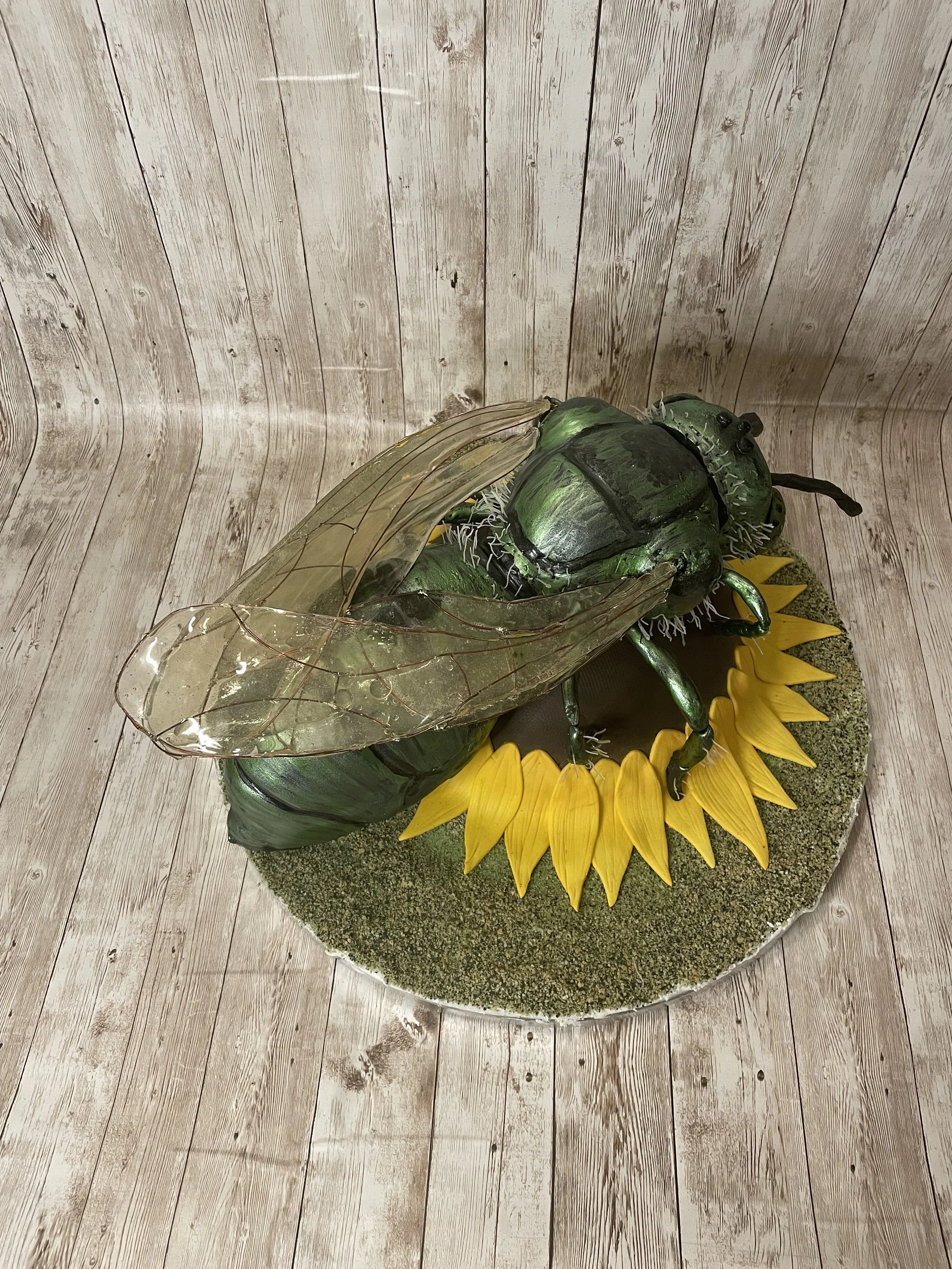 Large decorative cake green metallic sweat bee with transparent wings and black details, sitting on a sunflower with yellow petals and green center, on a round base with moss-like texture, against a wooden wall background.