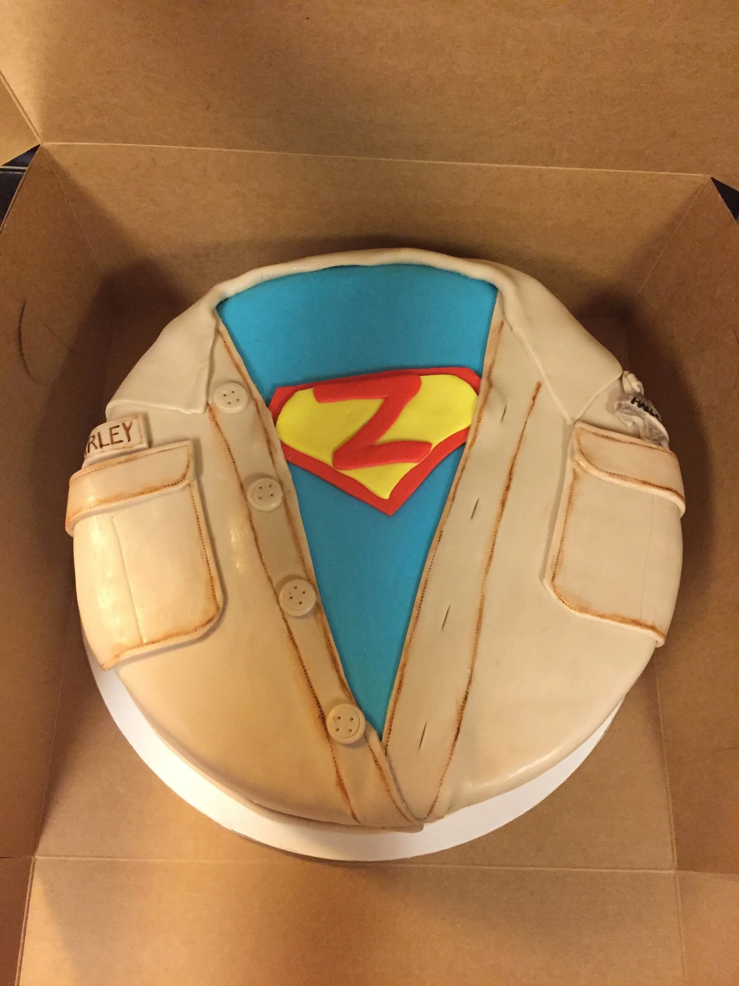 A round cake decorated as Superman's costume with a blue and yellow shield emblem and a beige shirt with buttons and pockets.