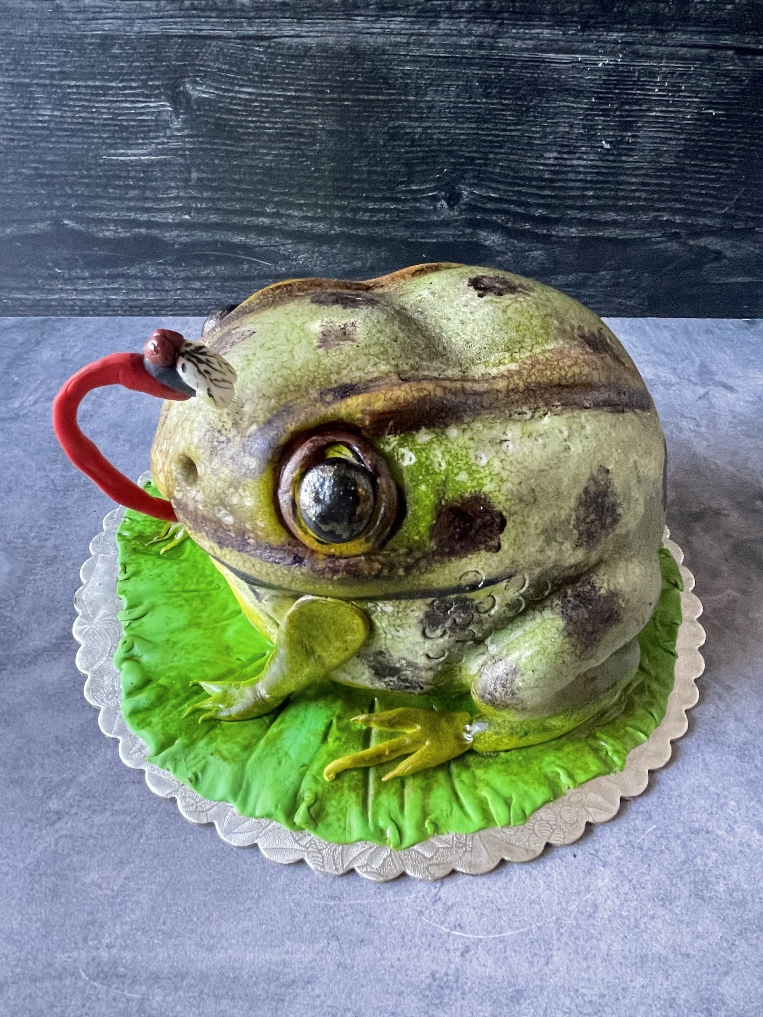 A cake designed to look like a frog, with detailed features including large eyes, a nose, and a body, sitting on a green leaf cake base with a decorative paper doily underneath.