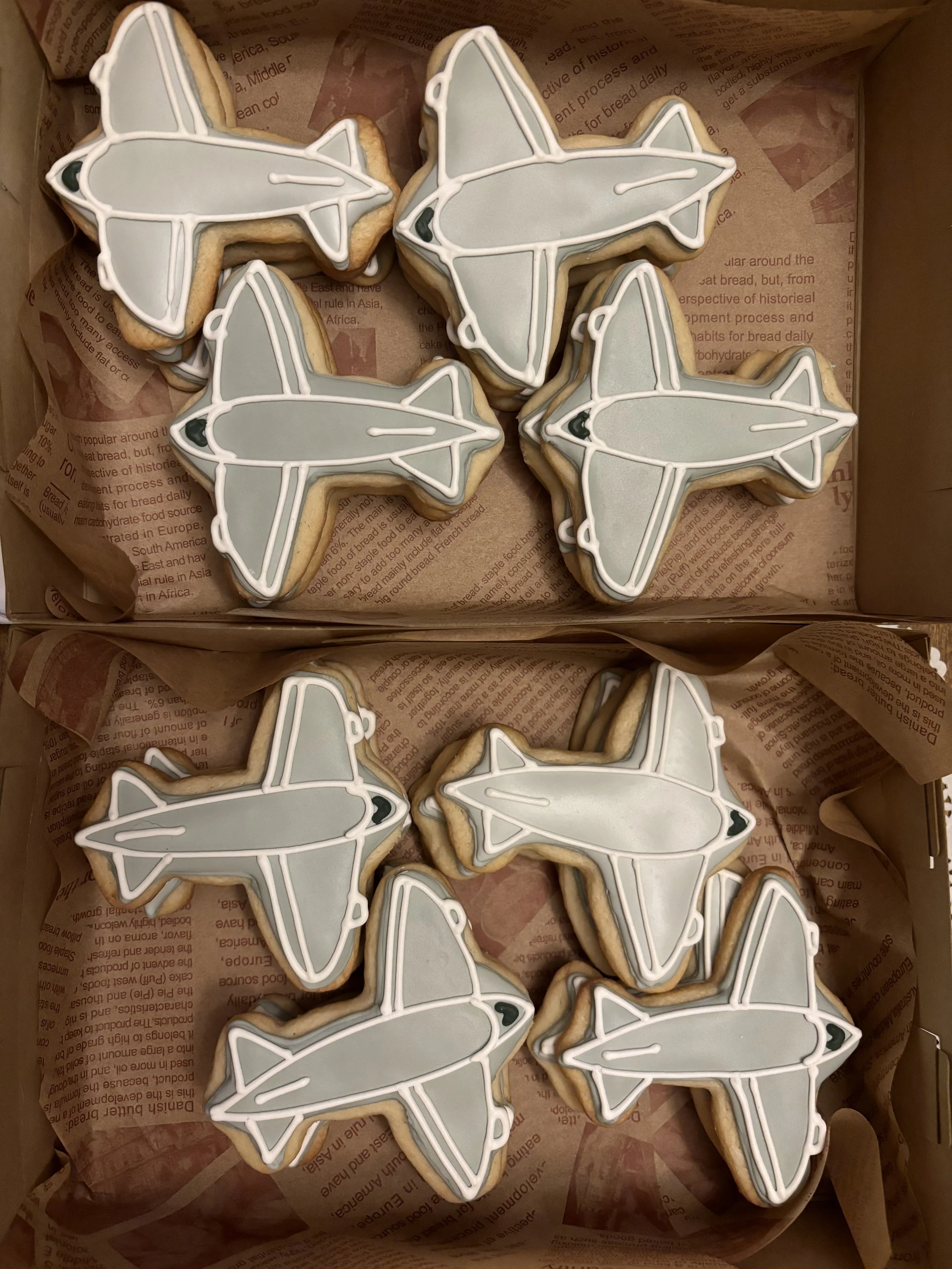 Cookies decorated with airplane designs in white, gray, and black icing arranged in a box lined with paper with text.