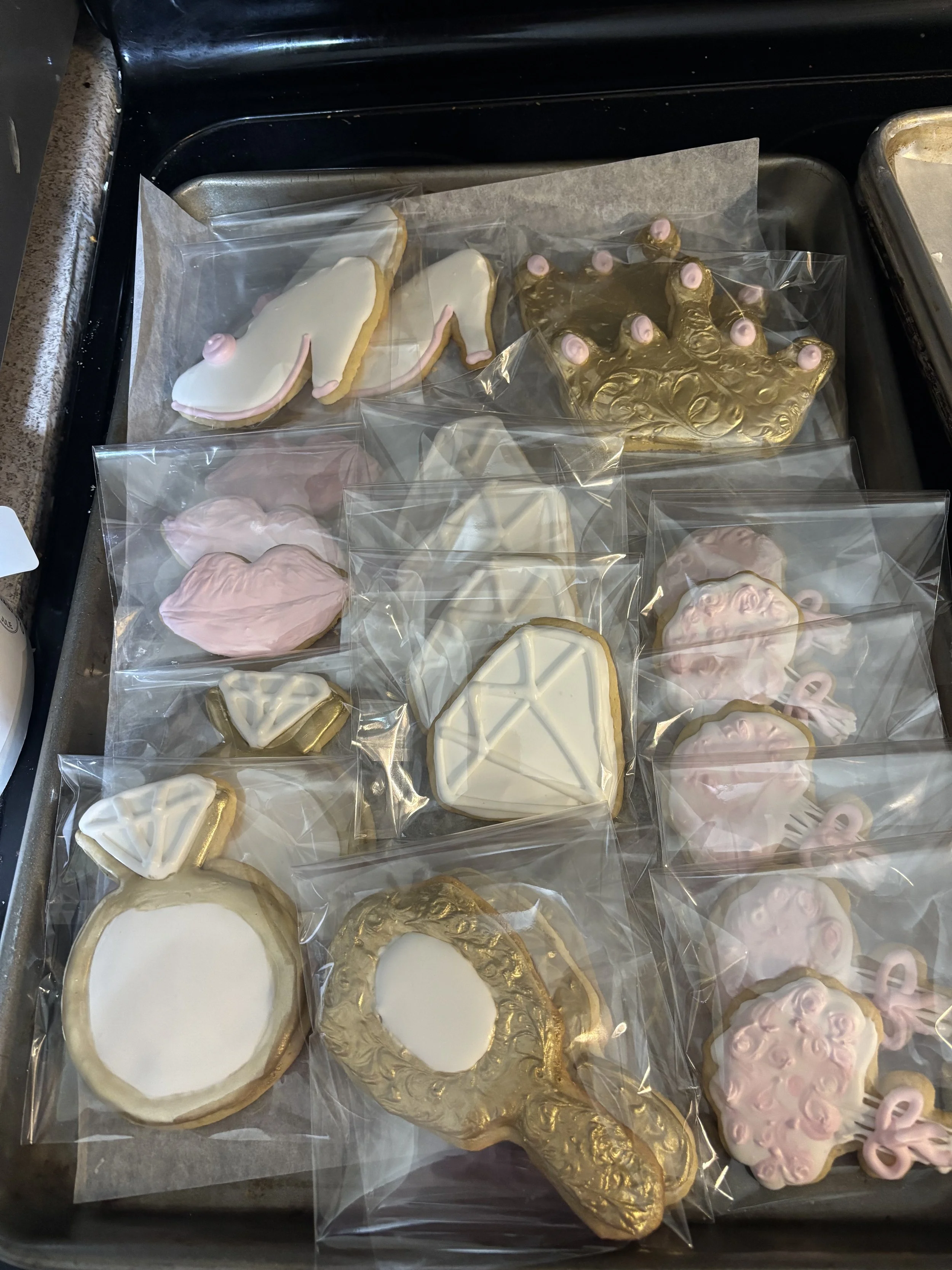 Assorted decorated cookies with pink, white, and gold icing and designs, individually wrapped in clear plastic.