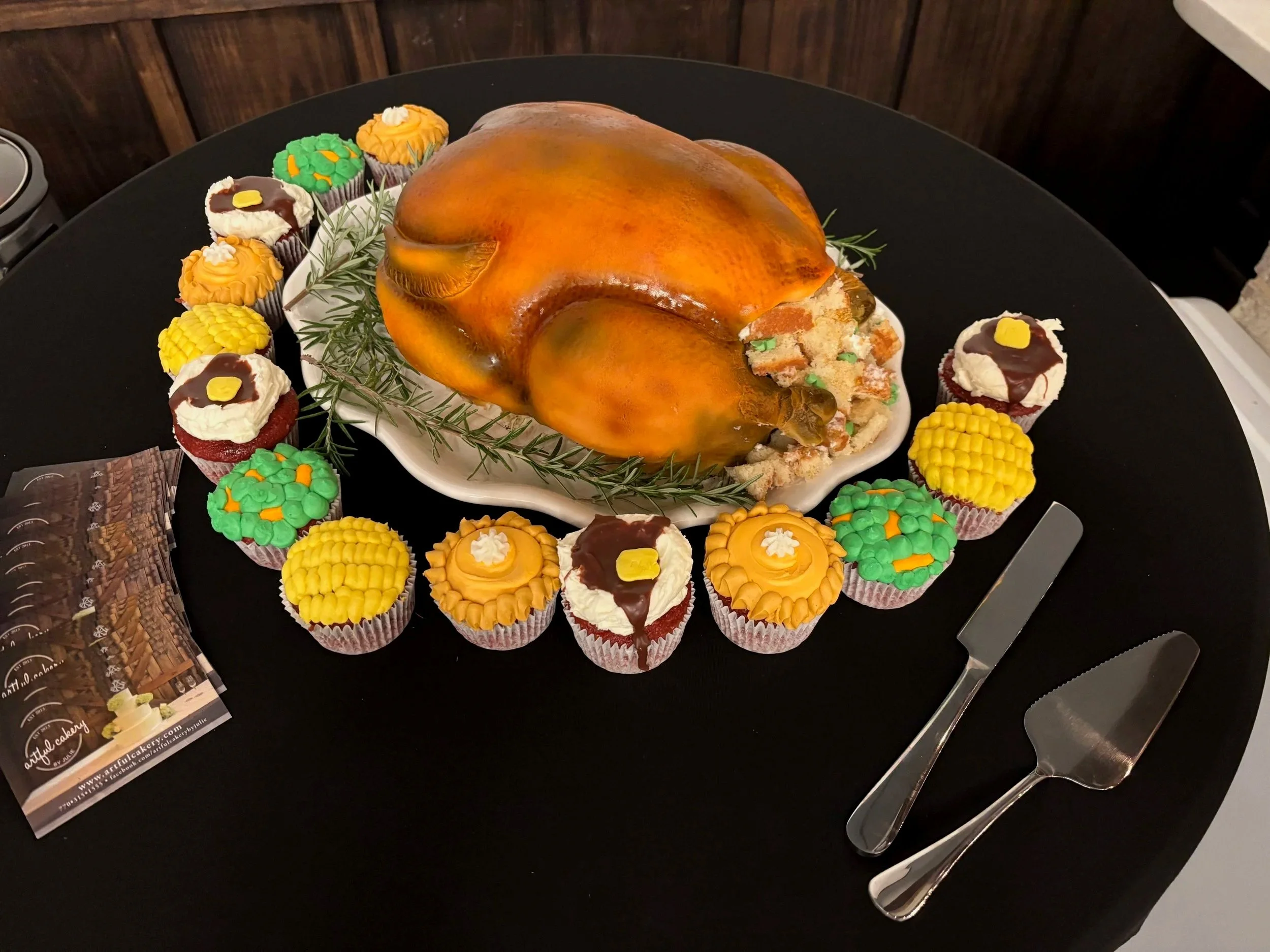 A life-like roasted turkey cake by Artful Cakery by Julie, complete with edible golden skin, herbs, and stuffing, surrounded by themed cupcakes decorated as classic Thanksgiving side dishes including mashed potatoes and gravy, corn on the cob, peas a