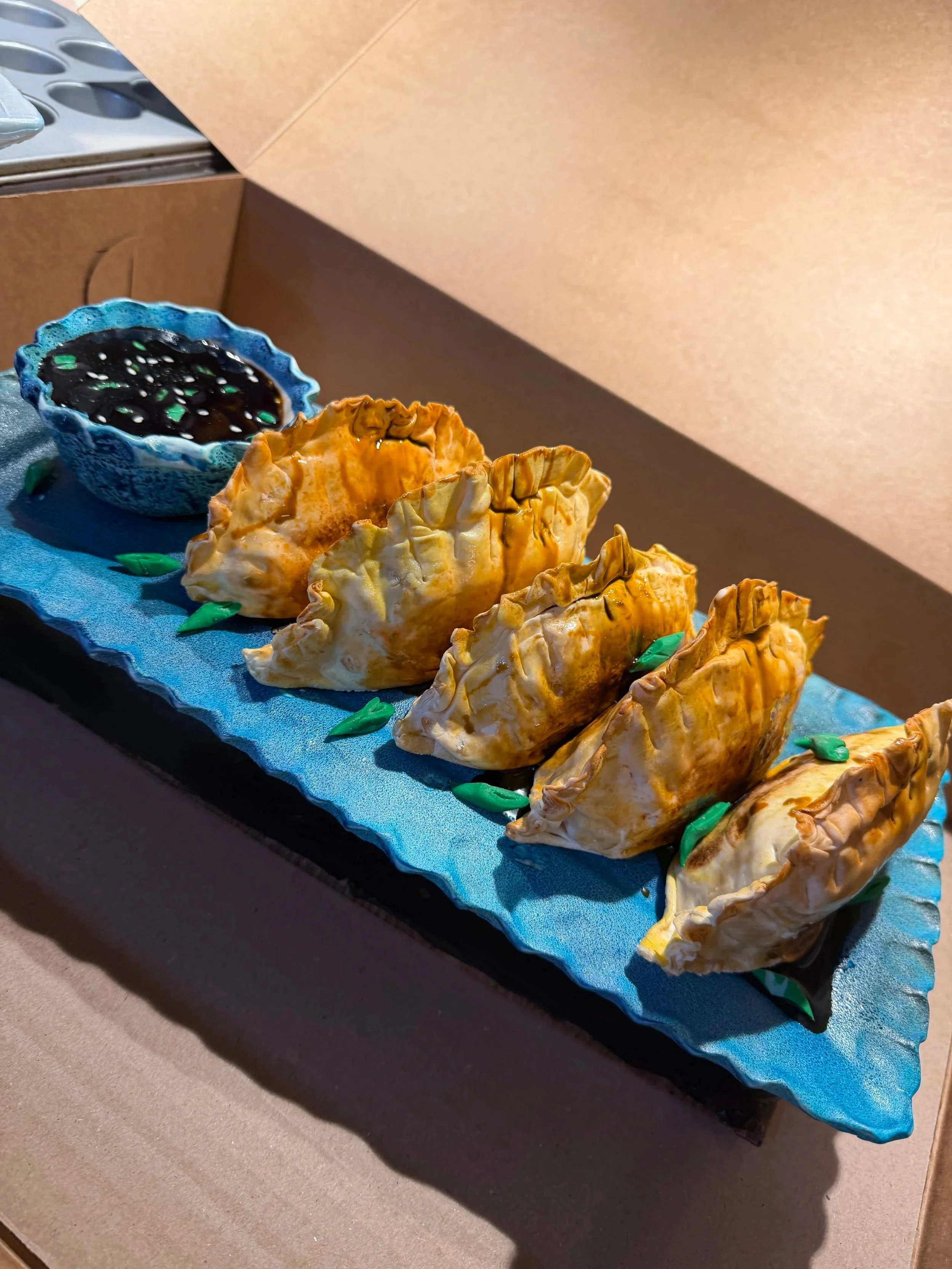 Realistic gyoza cake with five fried dumplings, edible blue serving platter, dipping sauce bowl, and green onion details.