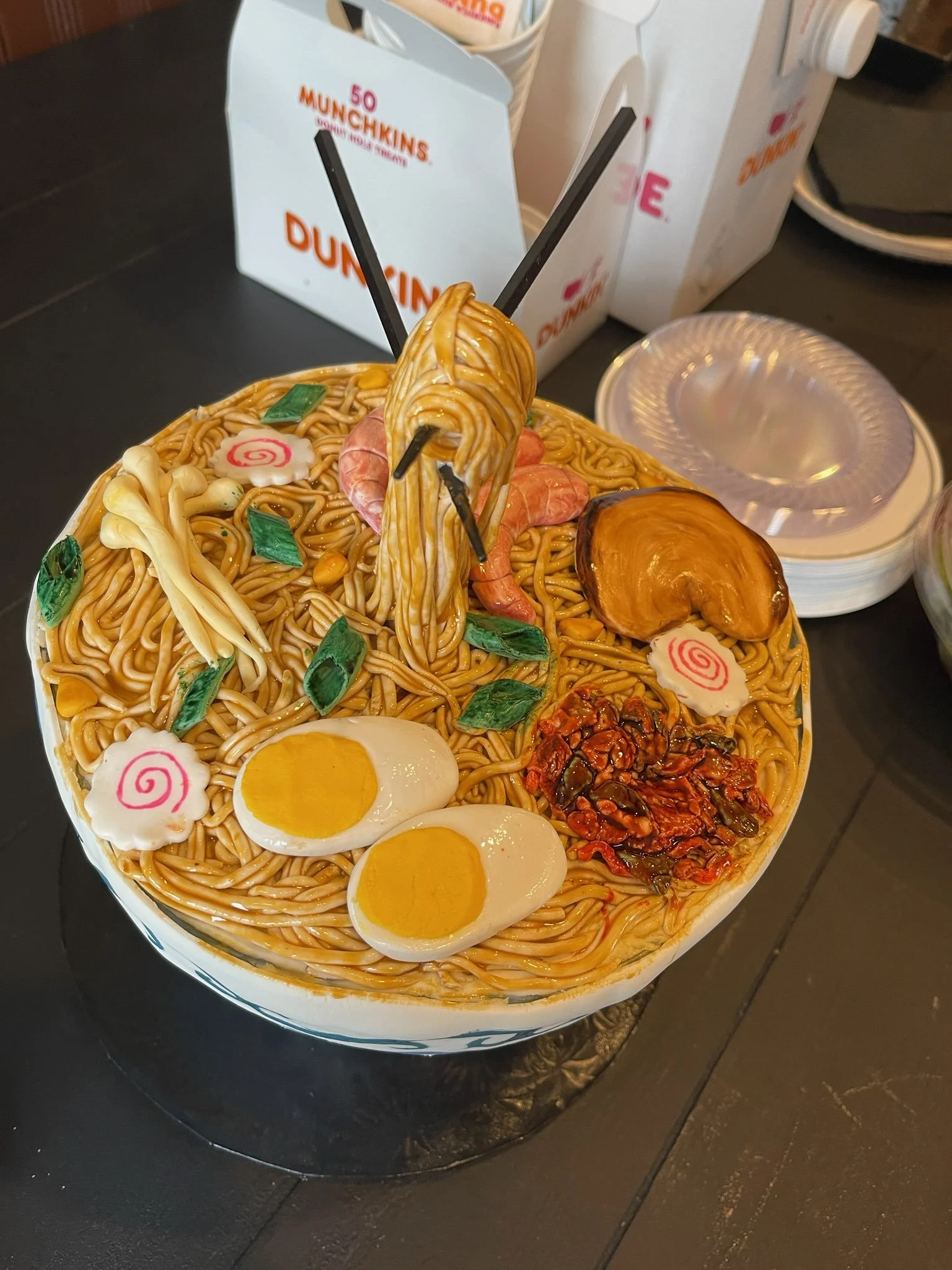 A cake sculpture made to look like a bowl of ramen, with noodles, boiled eggs, narutomaki fish cakes, green onions, mushrooms, shrimp, and other toppings, arranged to resemble a bowl of ramen.