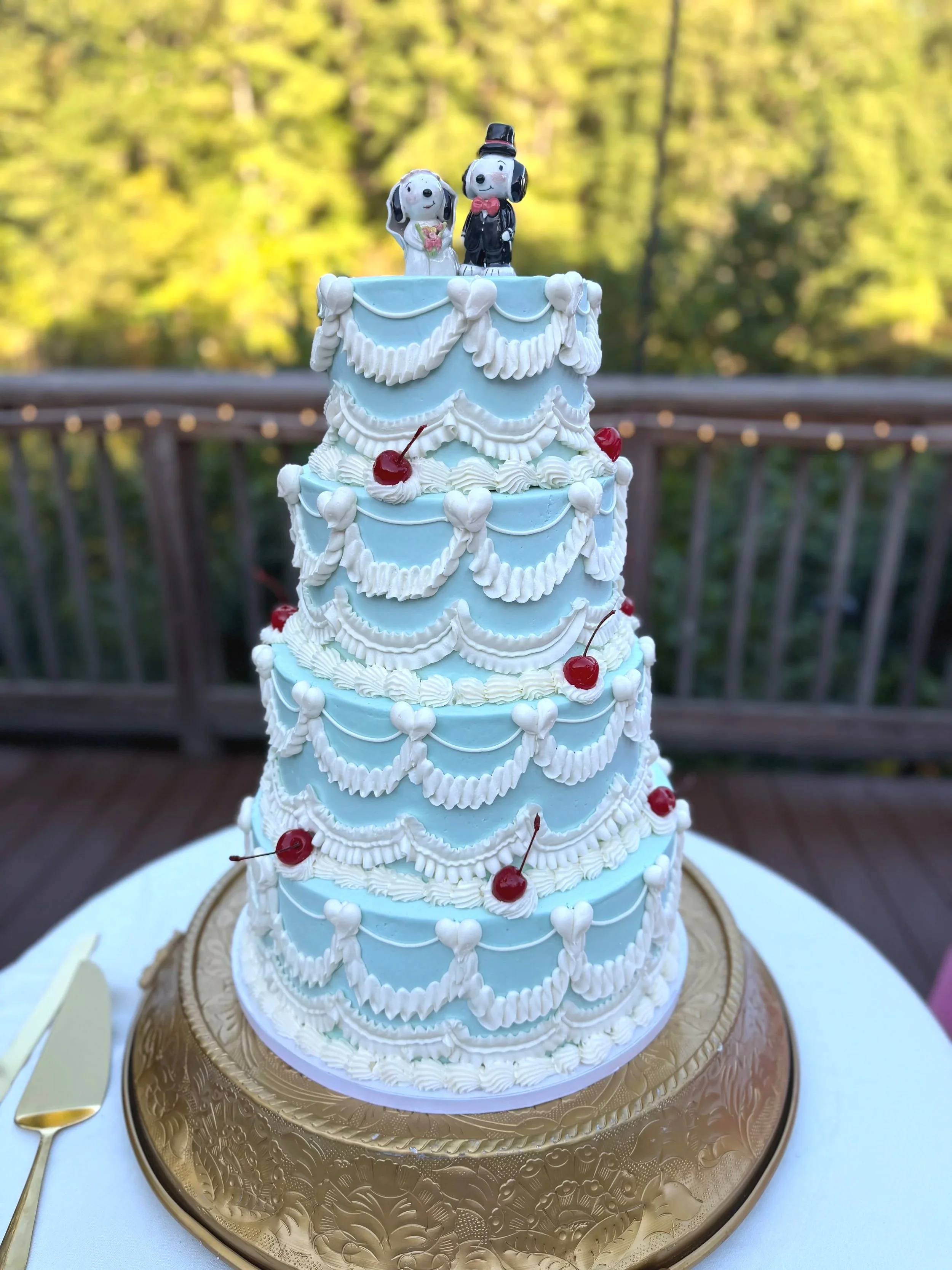 Four tiered vintage wedding cake frosted in blue with white piping and red cherries displayed at a reception in Chattahoochee Nature Center