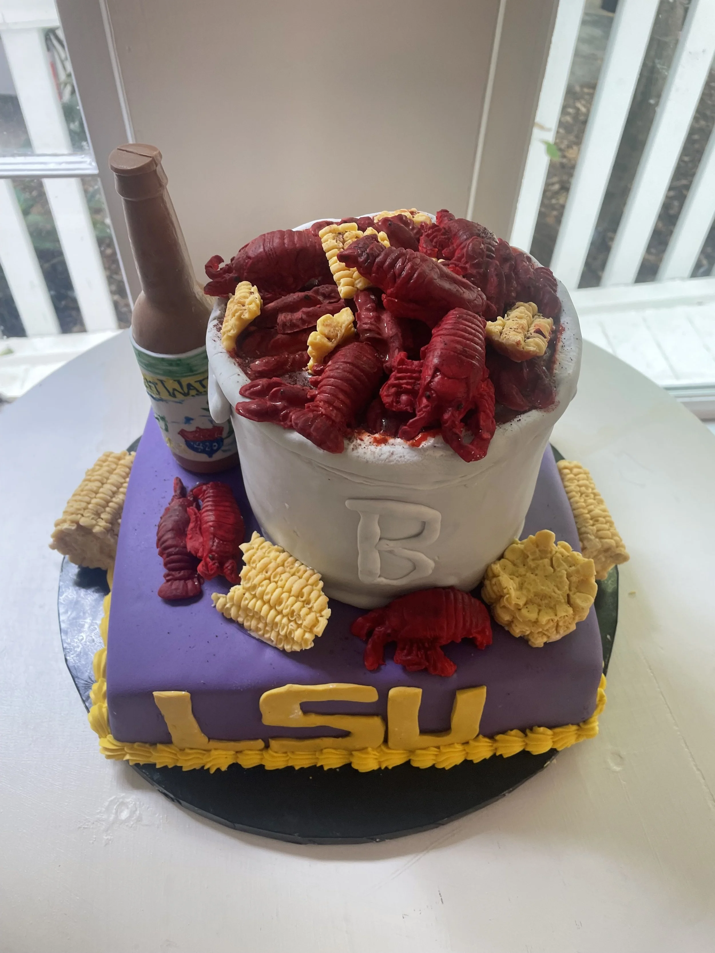 A cake with a purple base, yellow decorations, and a large pot on top filled with red crawfish and yellow corn, with a beer bottle in the background.