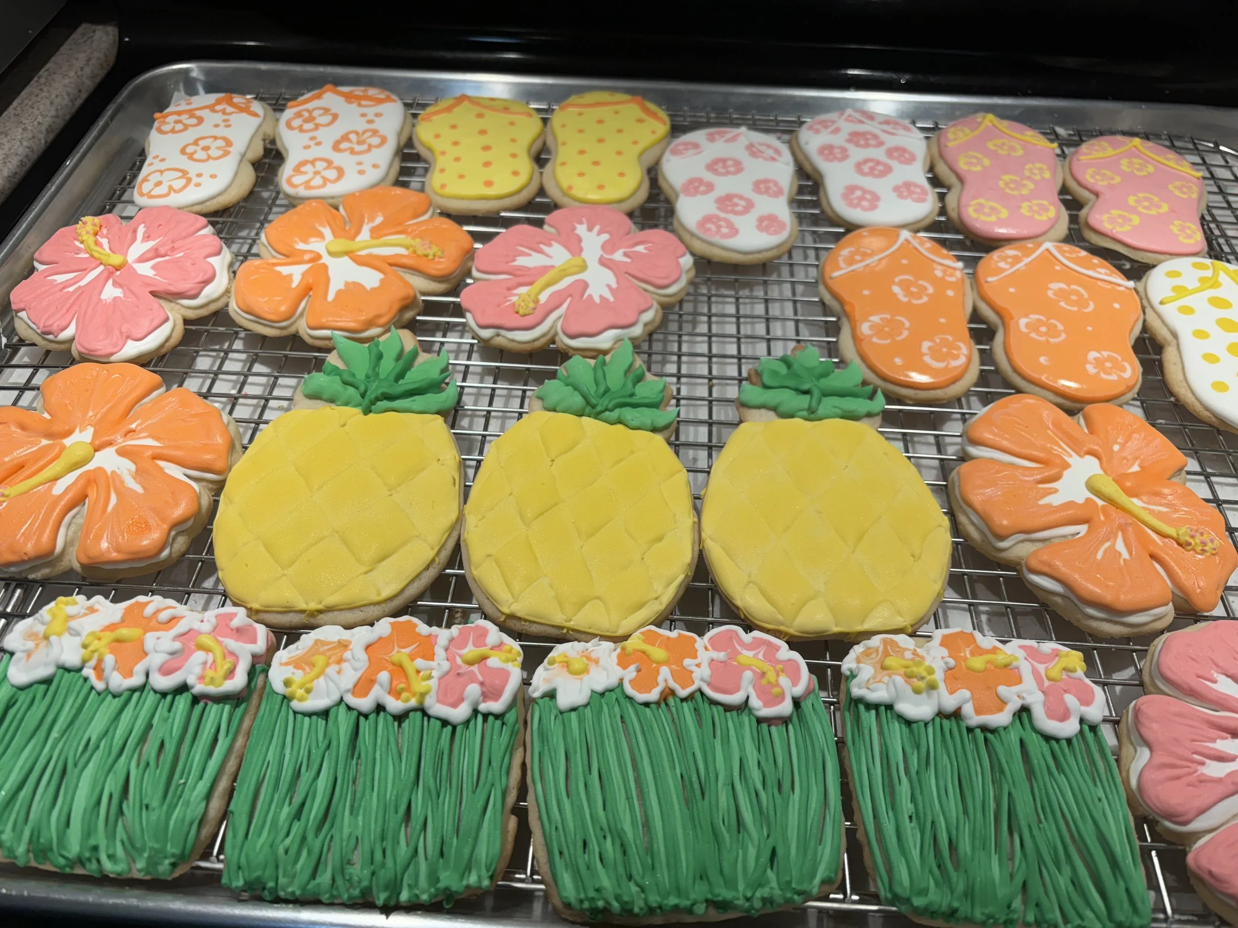 Cookies decorated as tropical flowers, pineapples, and grass, arranged on a cooling rack.