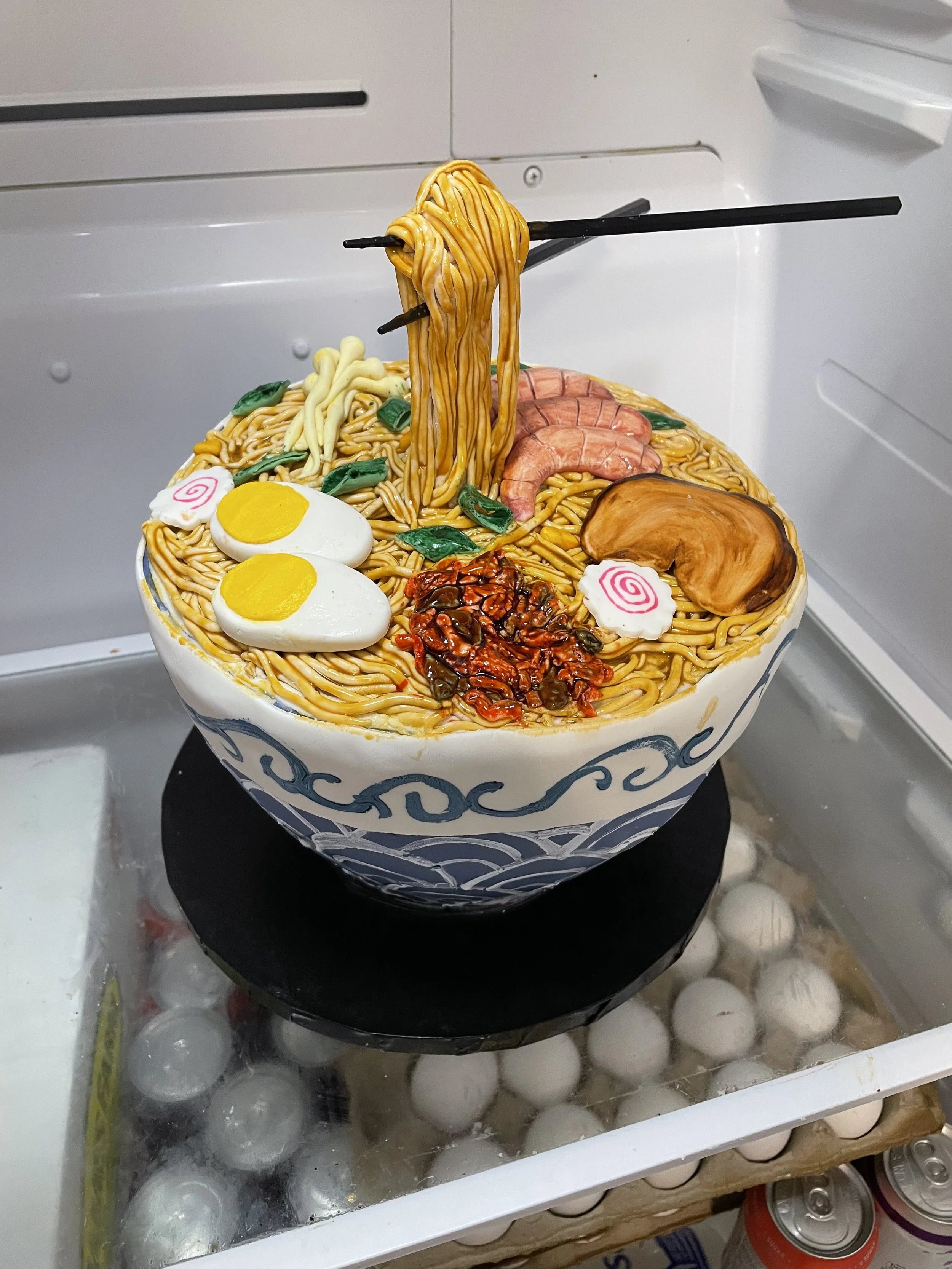 A cake designed to look like a bowl of ramen, with ramen noodles, eggs, shrimp, pork, green onions, and chili flakes, all made from edible materials.