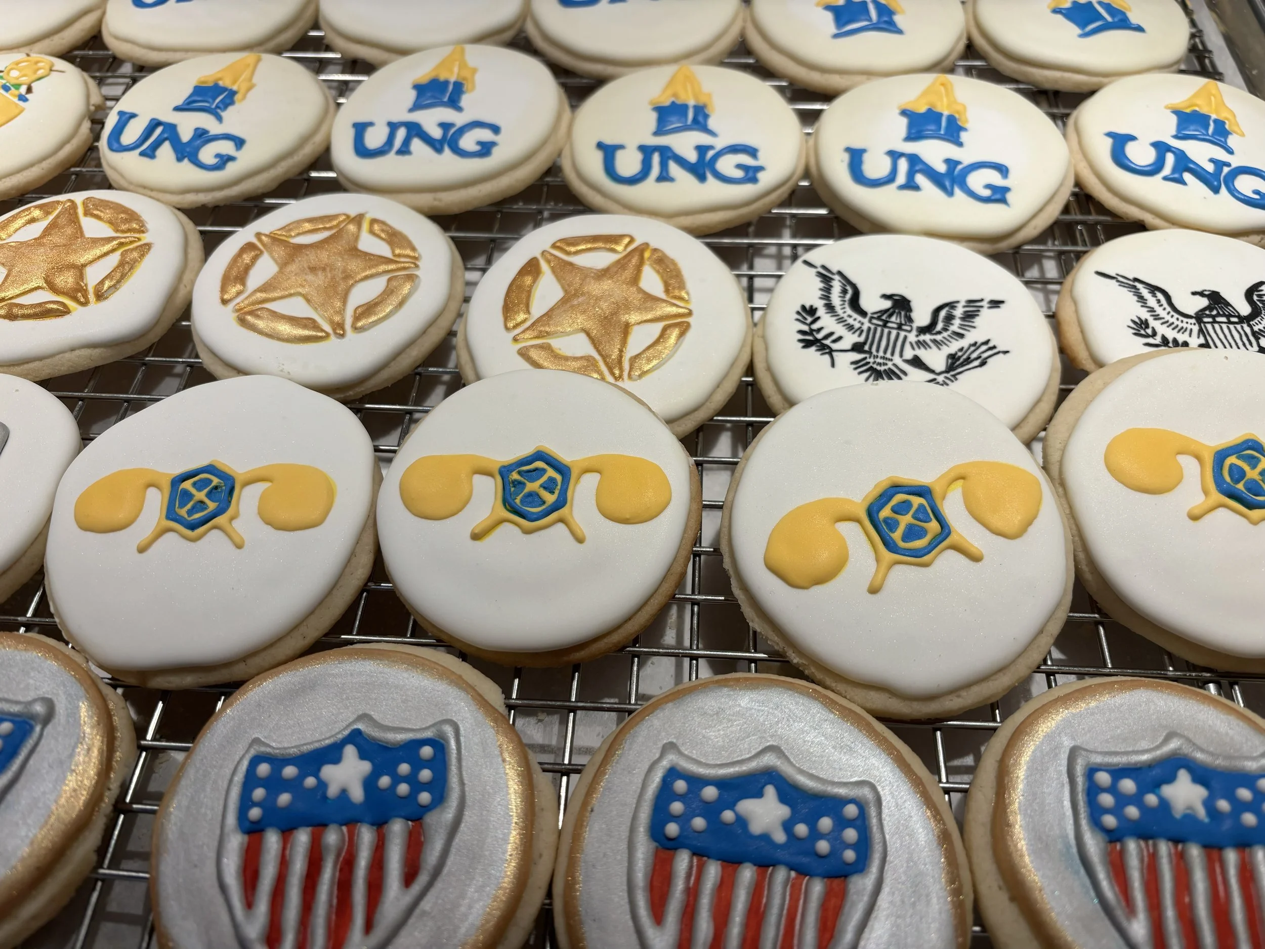Decorated cookies with patriotic logos, including 'UNG' with a blue and yellow logo, a gold star, and a black eagle emblem, arranged on a cooling rack.
