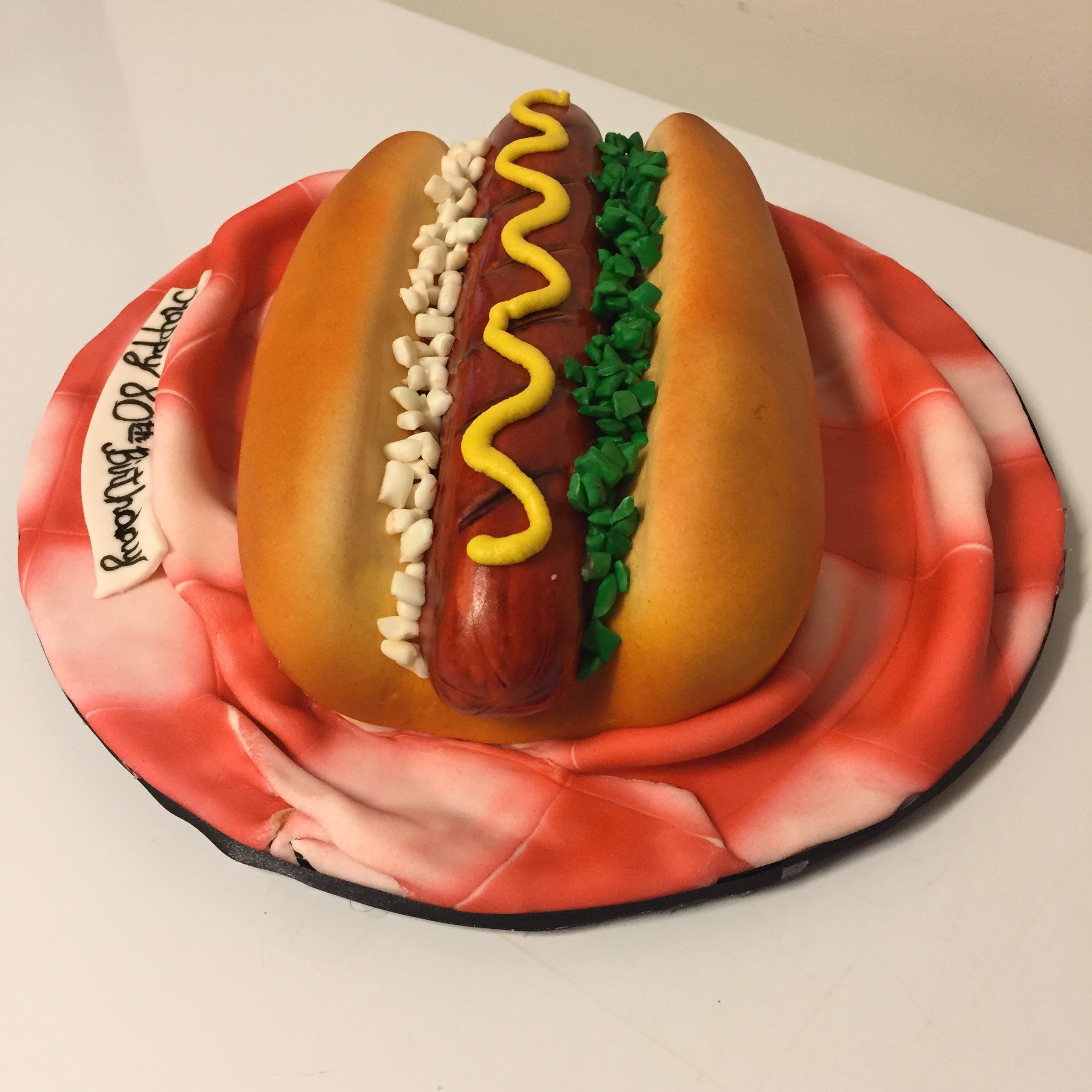 Cake designed to look like a hot dog in a bun with mustard, relish, and onions, on a patterned napkin