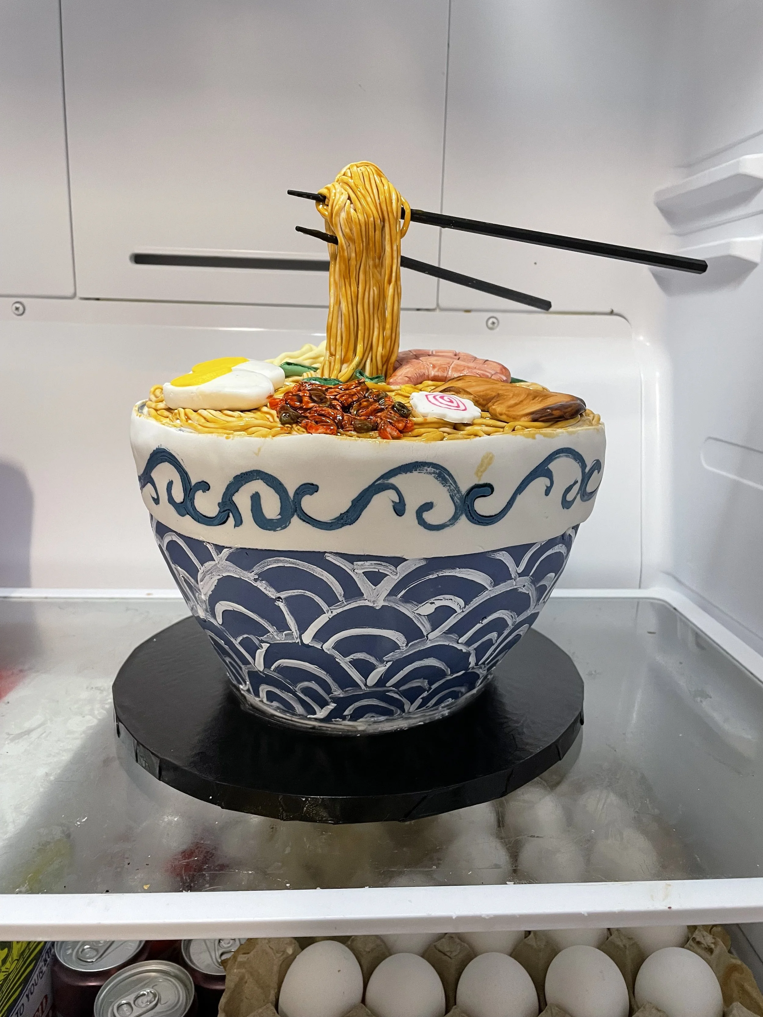 A decorative cake in the shape of a bowl of ramen with noodles, eggs, meat, and toppings, with chopsticks lifting noodles.