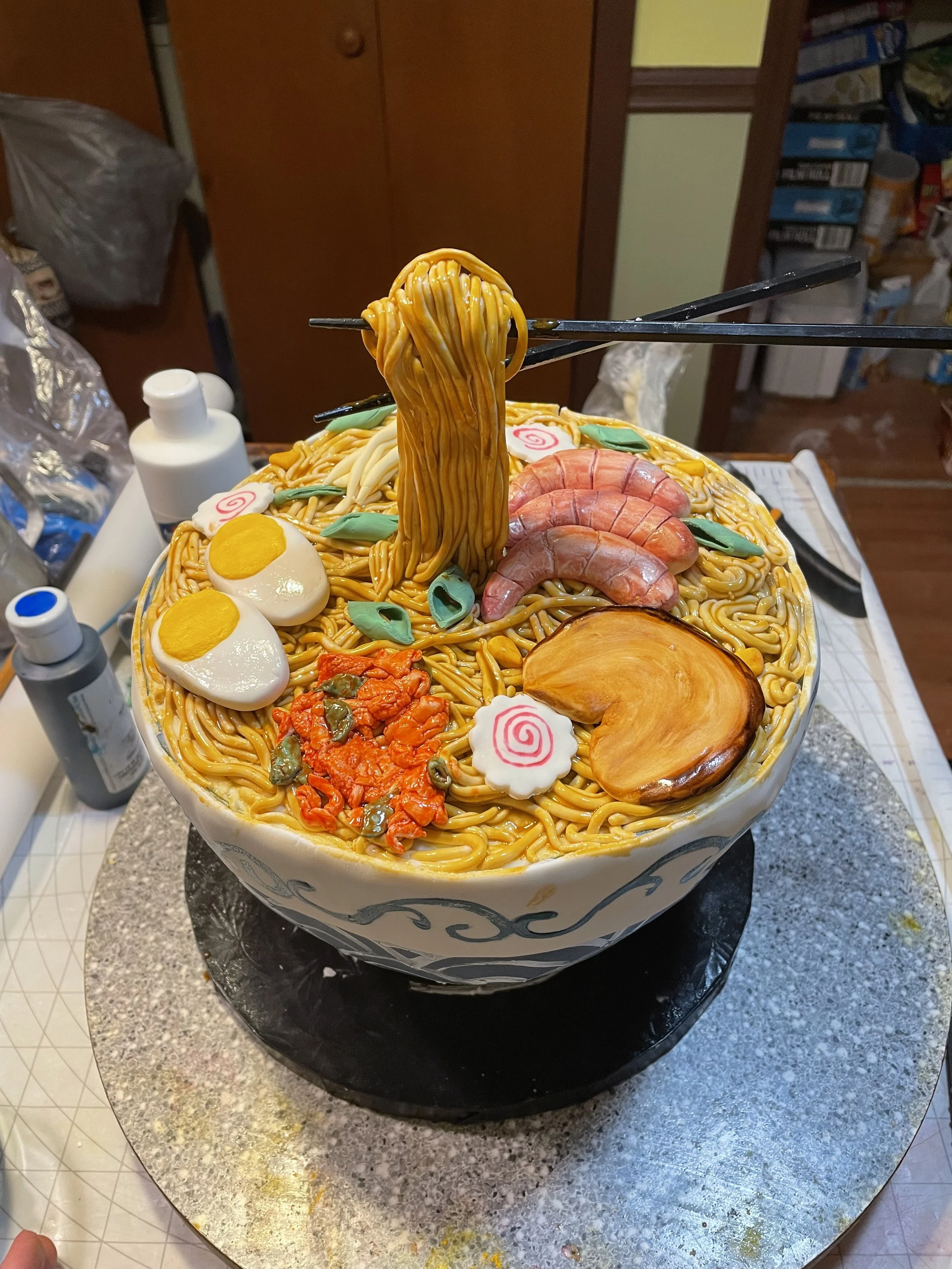 A cake decorated to look like a bowl of ramen noodles with toppings such as boiled eggs, shrimp, pork, narutomaki, and chili paste, with chopsticks lifting some noodles.